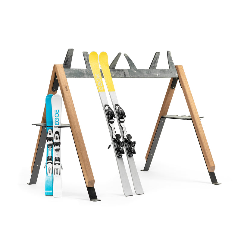 The Egoe Cabra Ski and Bike Rack is a multifunctional outdoor A-frame rack made from wood and metal. It holds two pairs of skis, one blue/white and one silver/yellow, both with bindings. Featuring extra supports and hooks for gear, it keeps your equipment protected while also storing bicycles.