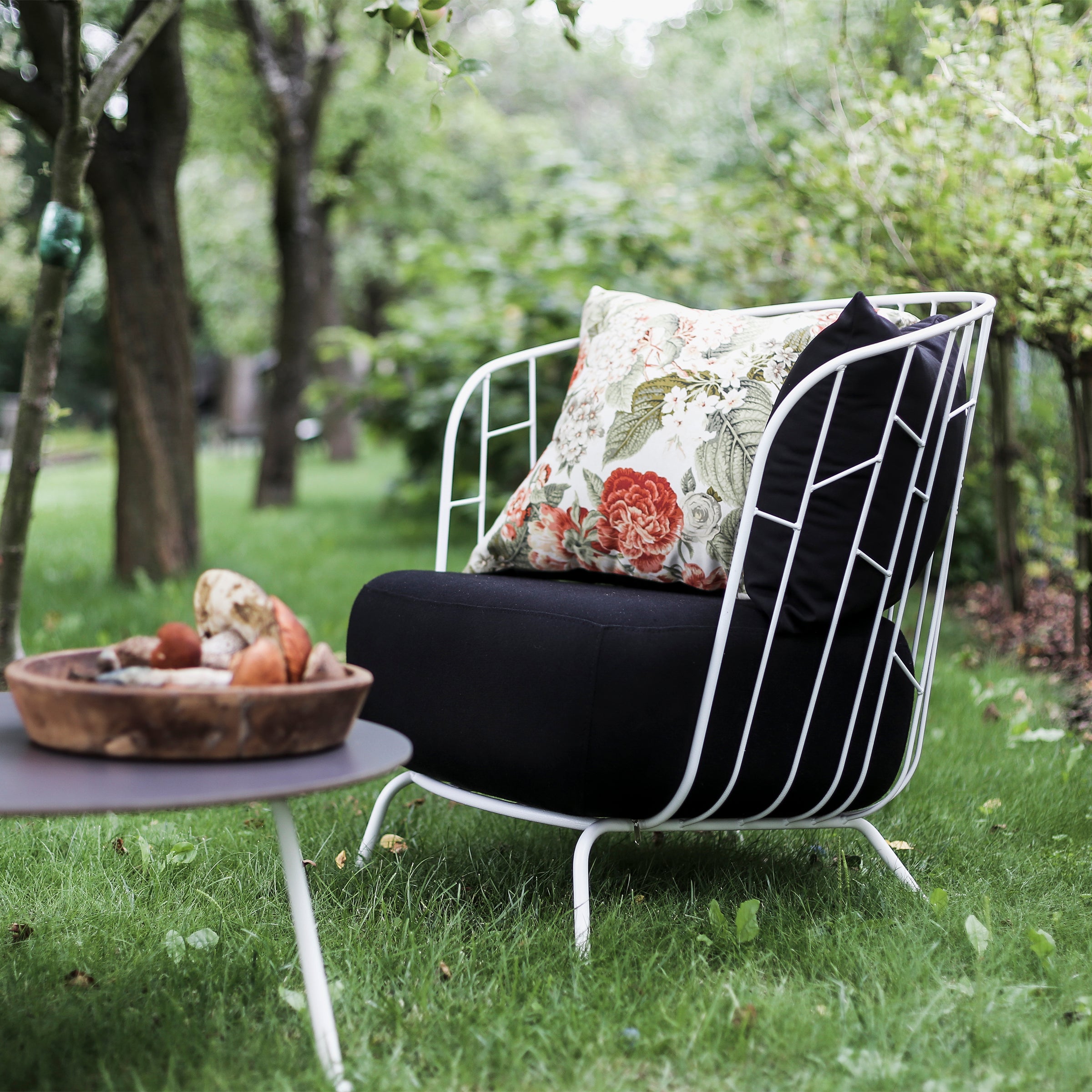The Egoe Maj Lounge Chair, with a black cushion and floral pillow, sits on lush grass. Beside it is a round zinc-coated steel table holding a wooden bowl filled with decorative stones, set against a backdrop of trees and greenery.