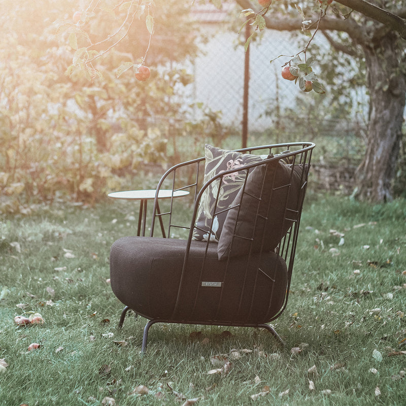 The Maj Lounge Chair by Egoe, complete with cushions and crafted from durable outdoor materials, sits on a grassy lawn strewn with fallen apples. A small side table complements the garden scene as sunlight filters through the tree branches above.