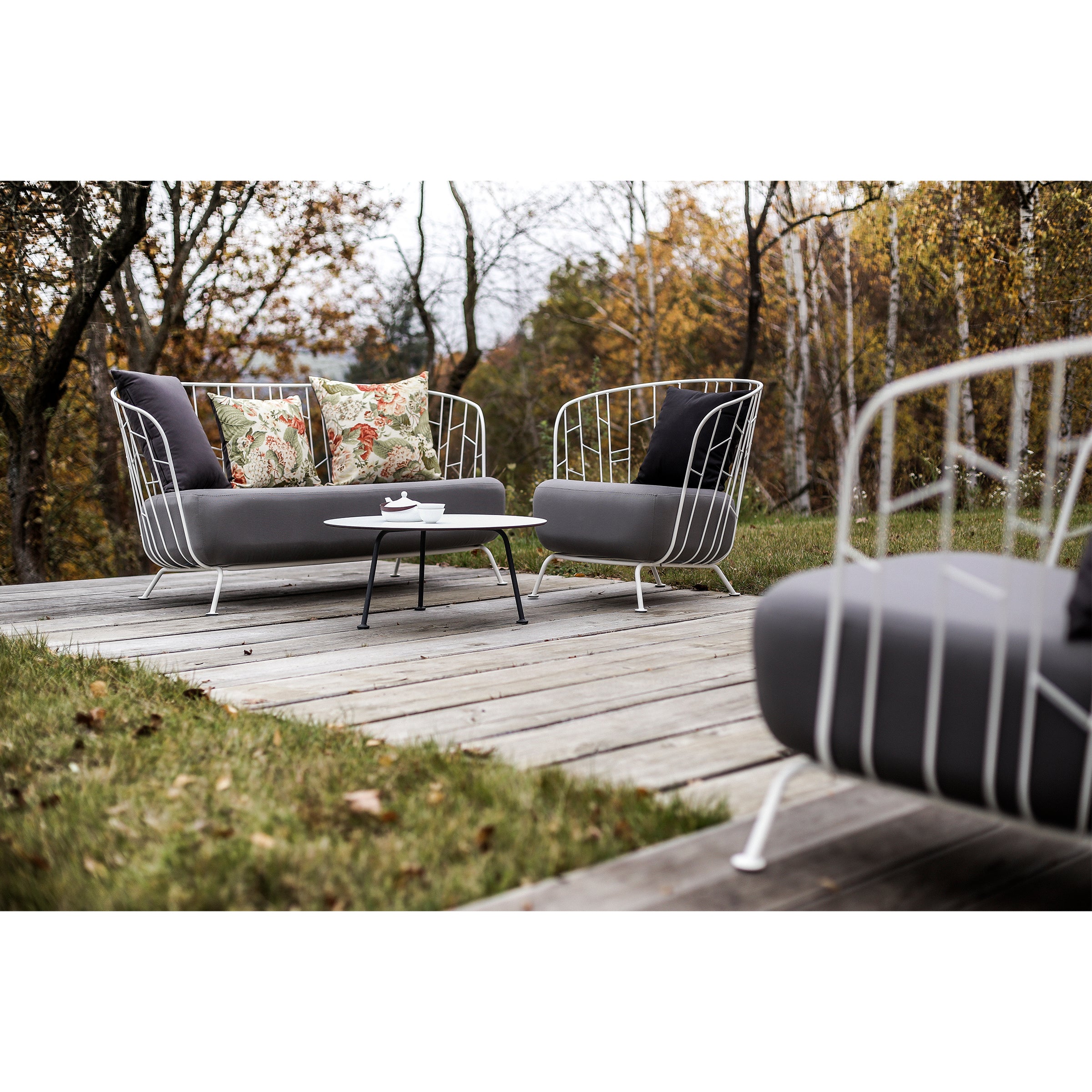 The outdoor patio features modern wire-frame chairs made from zinc-coated steel, adorned with gray cushions and floral pillows on a wooden deck. A small Egoe Maj Lounge Table stands between the chairs, complemented by trees with autumn foliage, highlighting the durable furnishings.