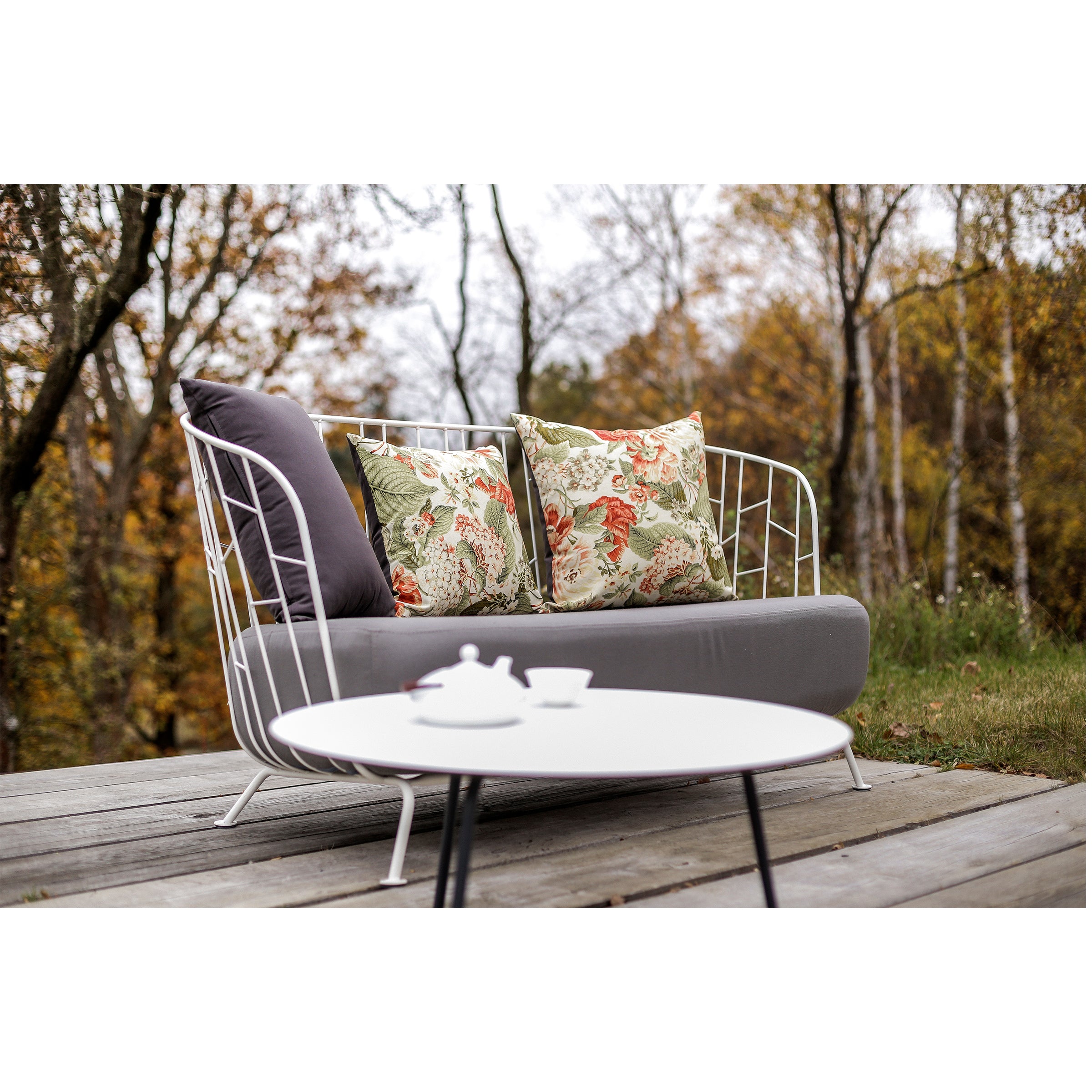 The outdoor seating area includes a gray cushioned sofa adorned with floral and plain pillows. An Egoe Maj Lounge Table, made from high-pressure laminate, holds a teapot and cup. Powder-coated steel accents add to the design while trees with autumn foliage create a serene atmosphere.