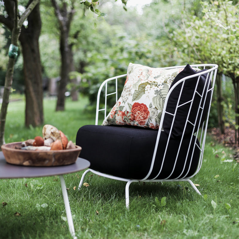 The Egoe Maj Lounge Chair, with a black cushion and floral pillow, sits on lush grass. Beside it is a round zinc-coated steel table holding a wooden bowl filled with decorative stones, set against a backdrop of trees and greenery.