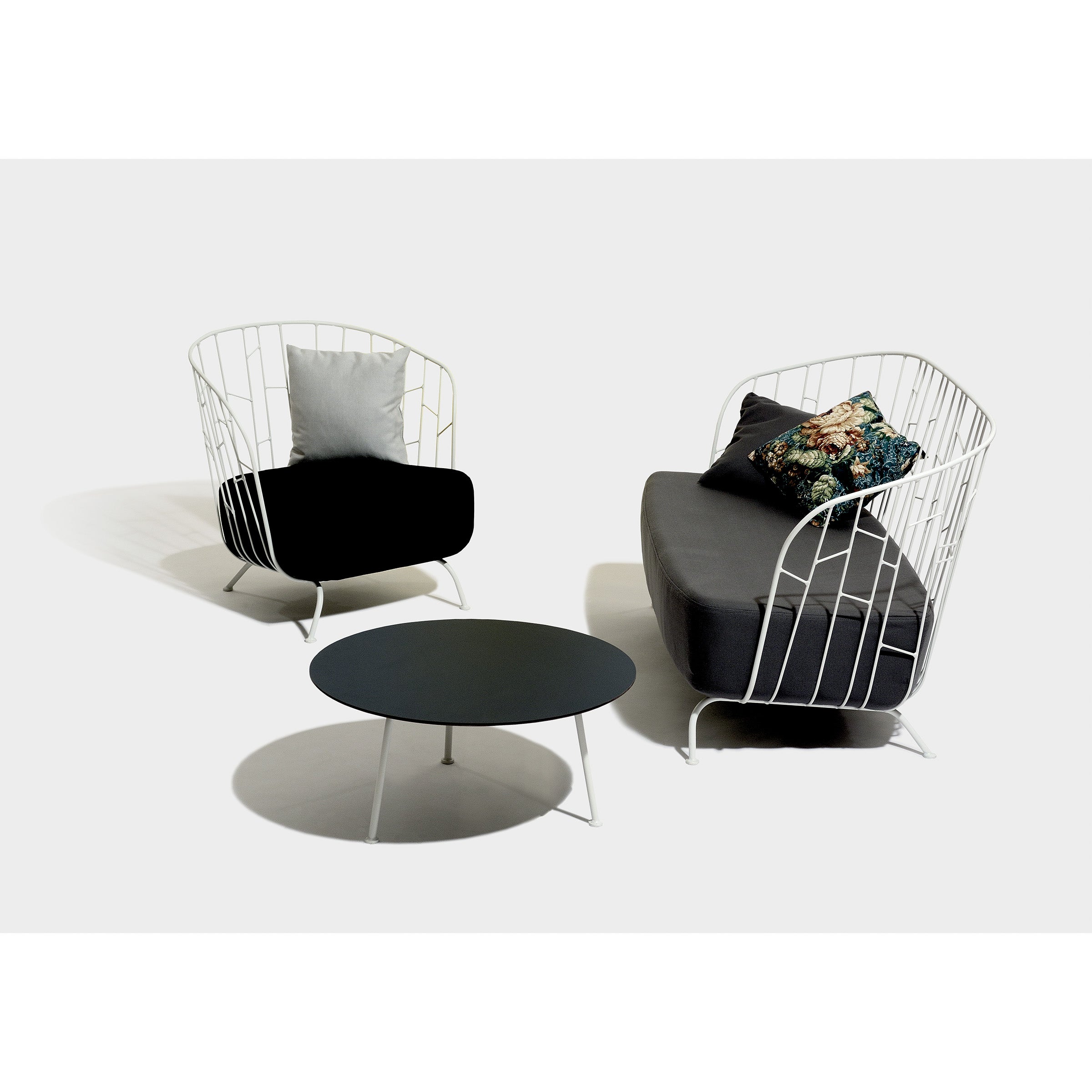 The Egoe Maj Lounge Chair is a white wireframe armchair crafted from zinc-coated steel, with a black cushion and gray pillow. Its paired with a matching loveseat featuring floral pillows and complemented by a round black coffee table, all set against a plain white background.