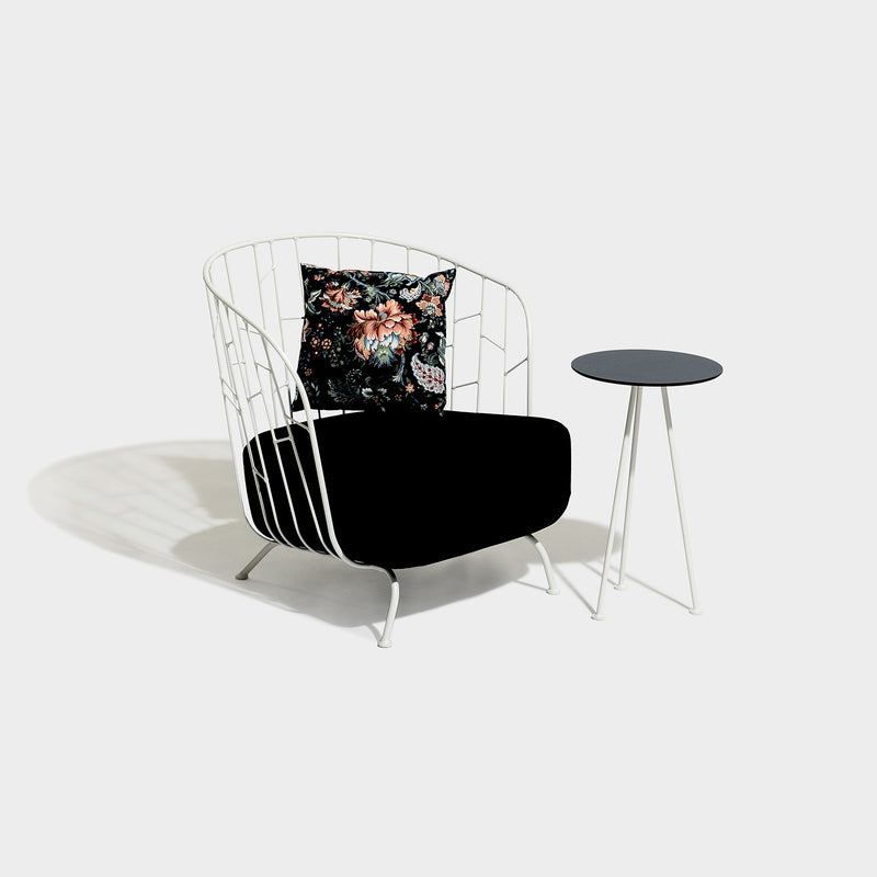 The Maj Lounge Chair by Egoe features a white zinc-coated steel frame, a black cushion, and a floral pillow. It sits beside a small round black side table against a plain gray background, ideal for highlighting the durability of outdoor materials.