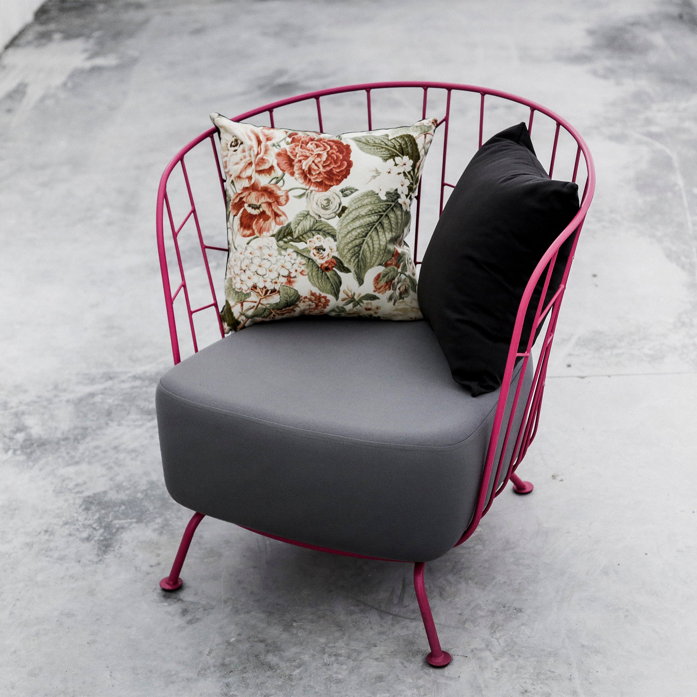 The Egoe Maj Lounge Chair boasts a vibrant pink wireframe made from zinc-coated steel. It includes a gray cushion with two pillows: one black and another floral-patterned, all elegantly placed on a concrete floor.