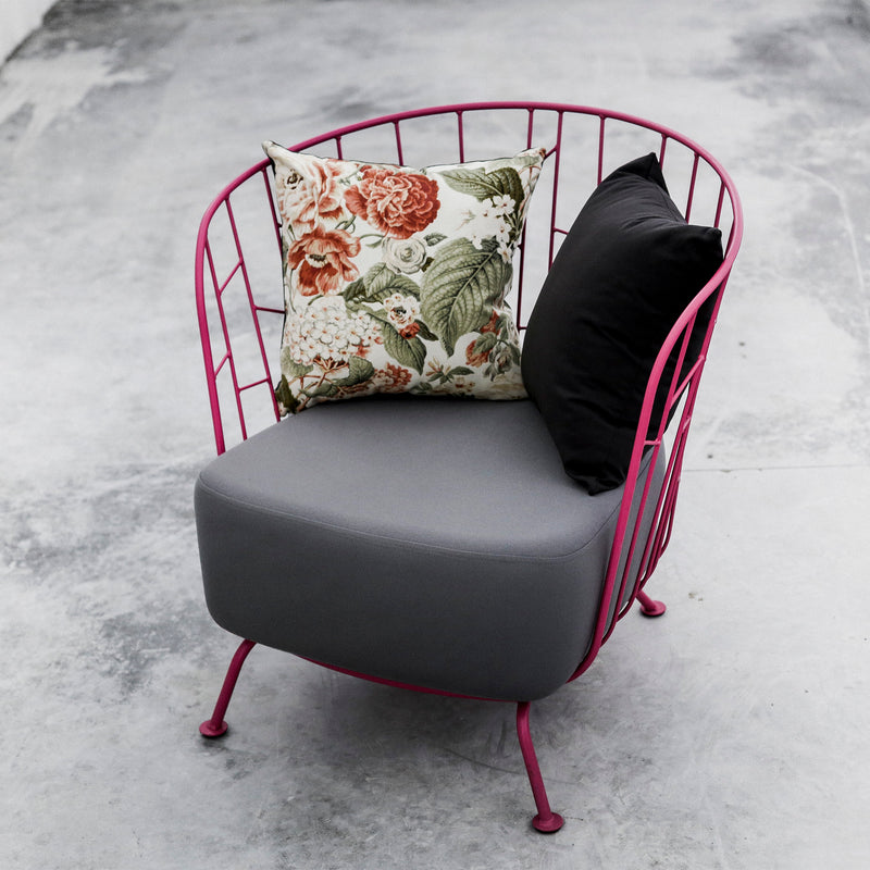 The Egoe Maj Lounge Chair boasts a vibrant pink wireframe made from zinc-coated steel. It includes a gray cushion with two pillows: one black and another floral-patterned, all elegantly placed on a concrete floor.
