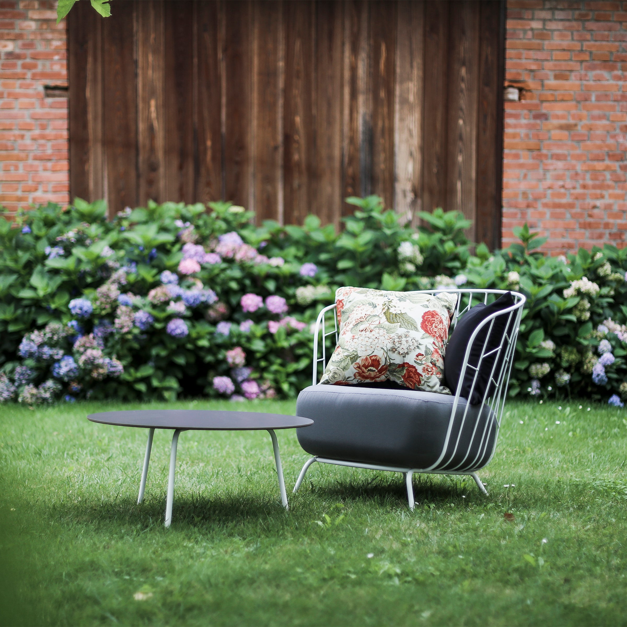 Outdoor seating area with Egoes Maj Lounge Chair featuring a floral cushion, beside a low black table on a well-manicured lawn. In the background, colorful hydrangea bushes and a rustic wooden and brick wall add charm to the scene.