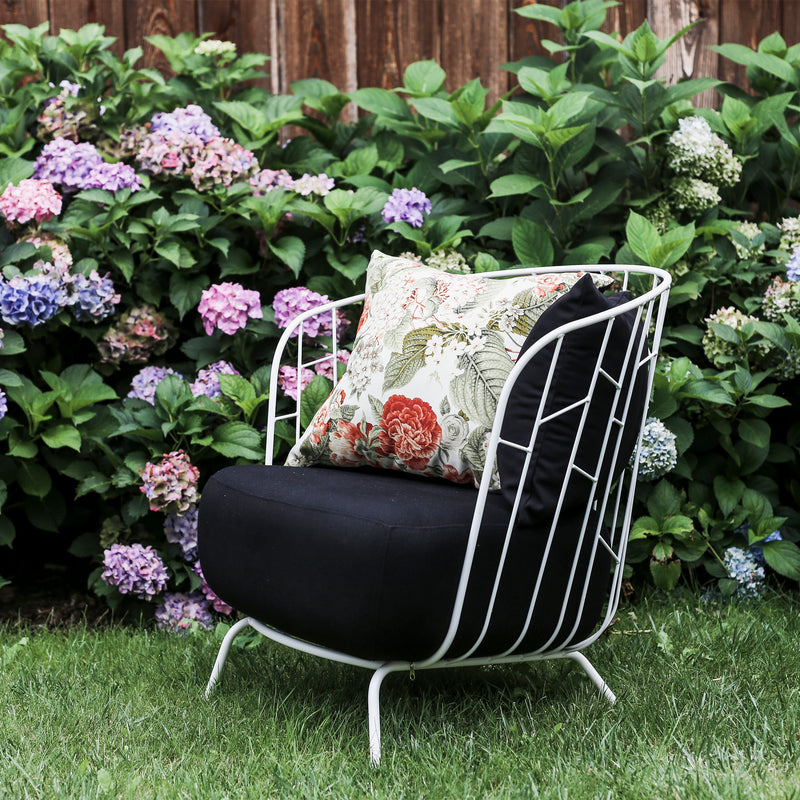 The Maj Lounge Chair by Egoe, with a zinc-coated steel frame, features a black cushion and a floral decorative pillow. Its placed on the grass in a garden, set against a wooden fence adorned with blooming hydrangeas showcasing pink, purple, and blue flowers.