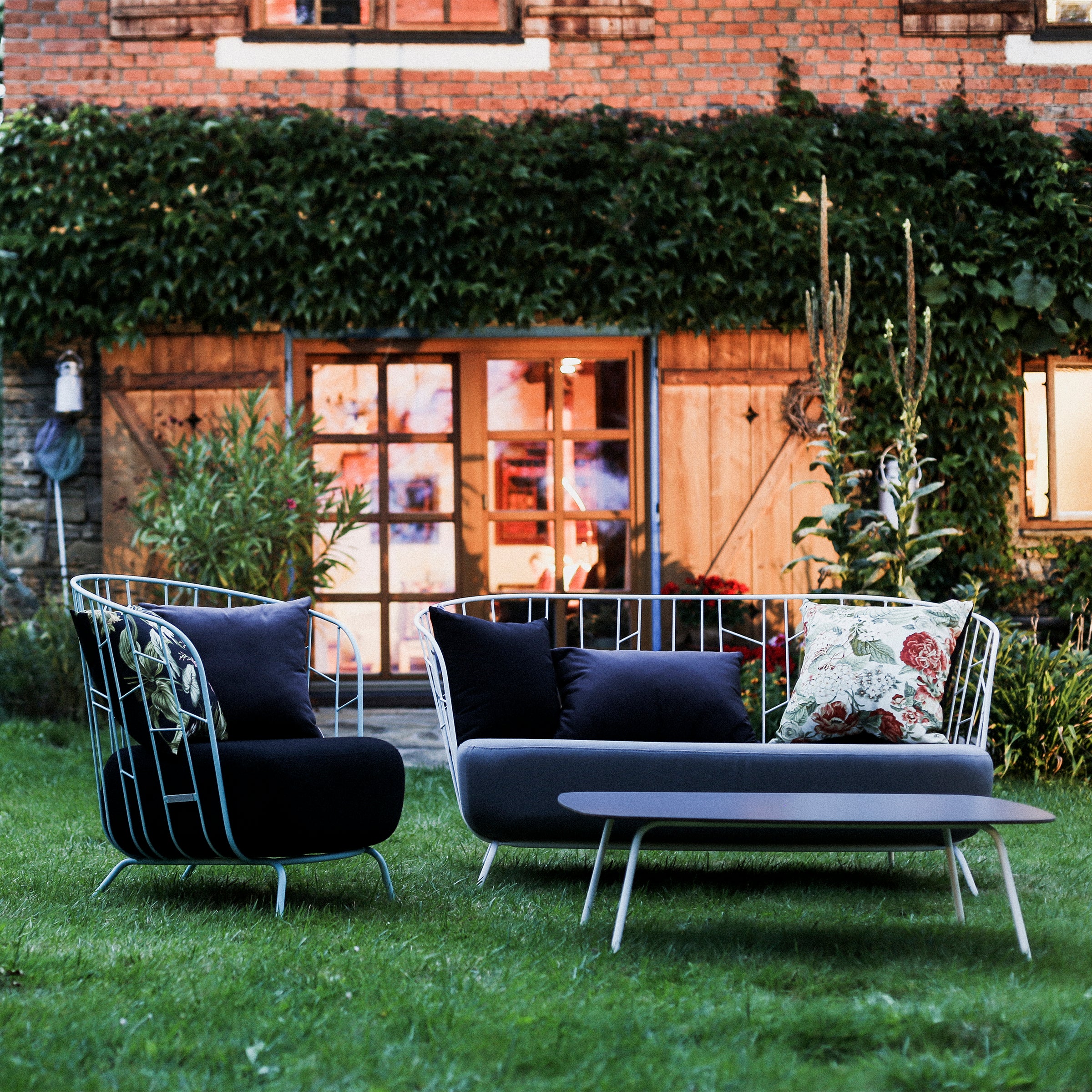 A lush green lawn accentuates Egoes Maj Lounge Table, positioned in front of a building with ivy-covered brick walls and large, illuminated windows. Featuring zinc-coated steel with a powder-coated finish, it includes a wire-frame loveseat and chair adorned with dark cushions.