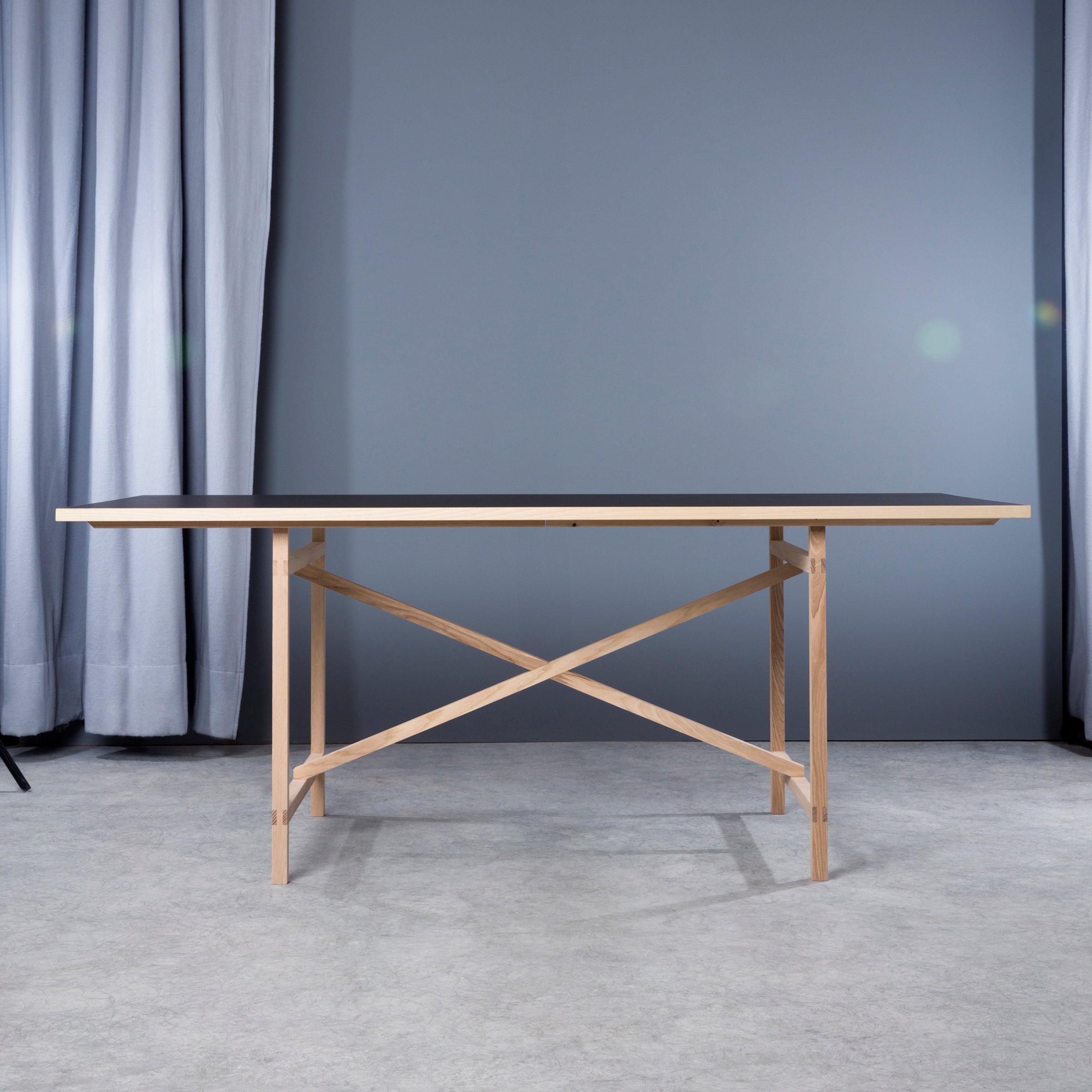 The Egon table by Moormann features a slim black top with a light wooden X-shaped base. Displayed in a minimalist room, it complements the gray walls and curtains, and the light gray floor highlights its elegant, metal-free design.