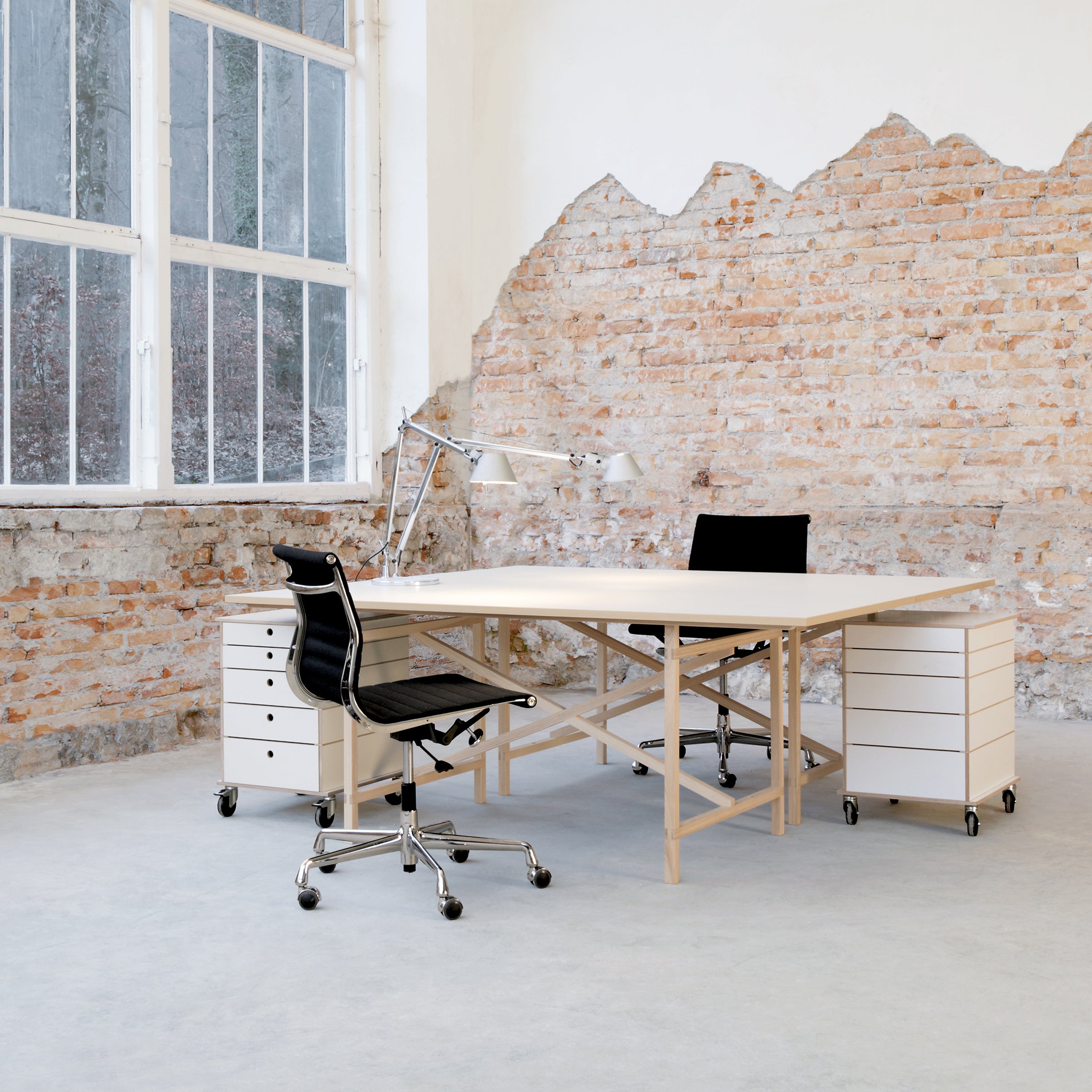 A minimalist office showcases Moormanns Egon wooden table with two black chairs, flanked by white wheeled drawer units. A large window and exposed brick wall provide an industrial feel, while a gray floor completes the space.