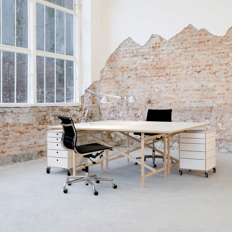 A minimalist office showcases Moormanns Egon wooden table with two black chairs, flanked by white wheeled drawer units. A large window and exposed brick wall provide an industrial feel, while a gray floor completes the space.