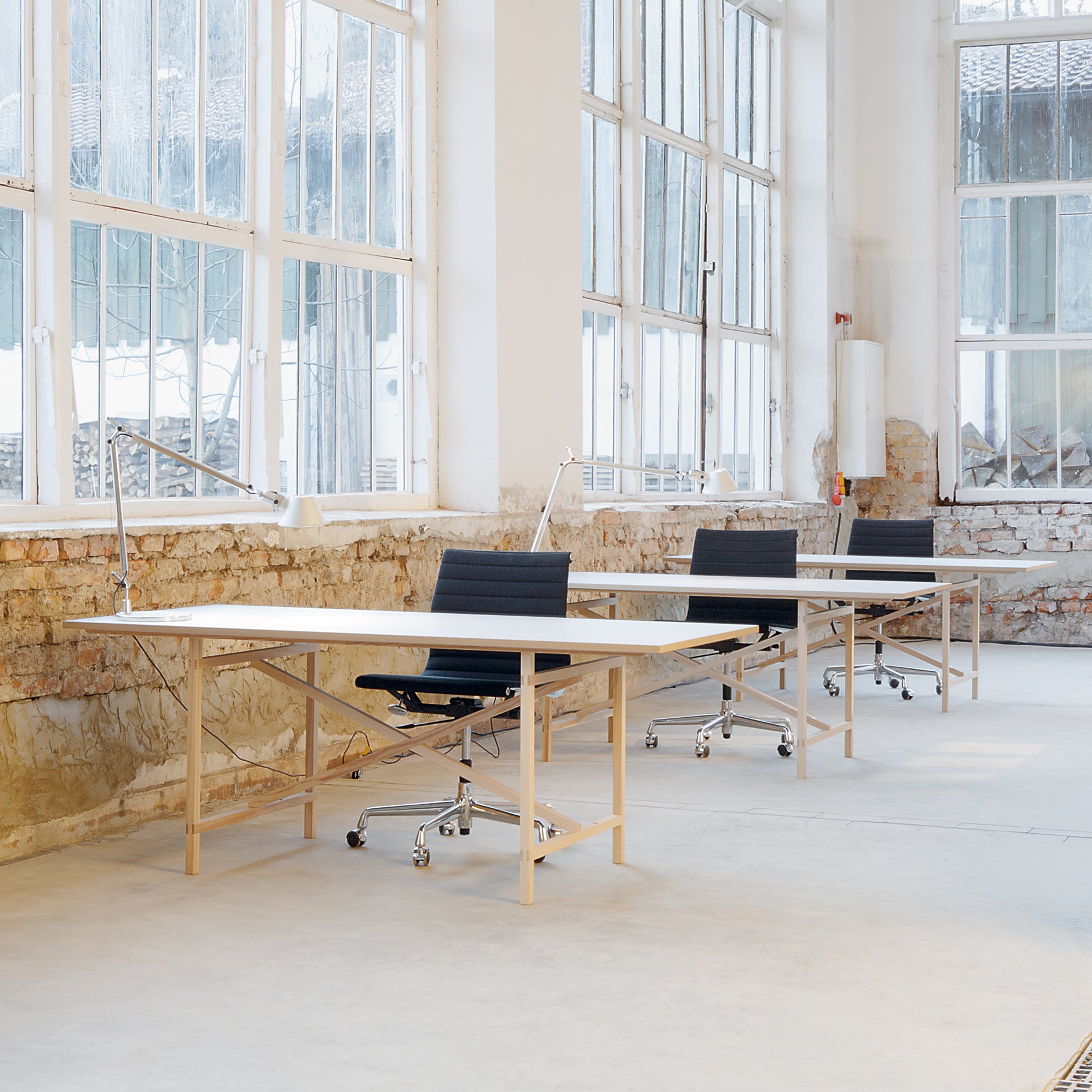 A minimalist office with large windows reveals rustic brick walls. The room features four Egon light wooden desks by Moormann, each paired with a black office chair and a white desk lamp. The setting is airy and modern, reflecting Franz Volhards simplicity ethos.
