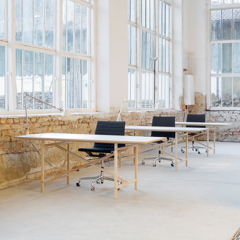 A minimalist office with large windows reveals rustic brick walls. The room features four Egon light wooden desks by Moormann, each paired with a black office chair and a white desk lamp. The setting is airy and modern, reflecting Franz Volhards simplicity ethos.