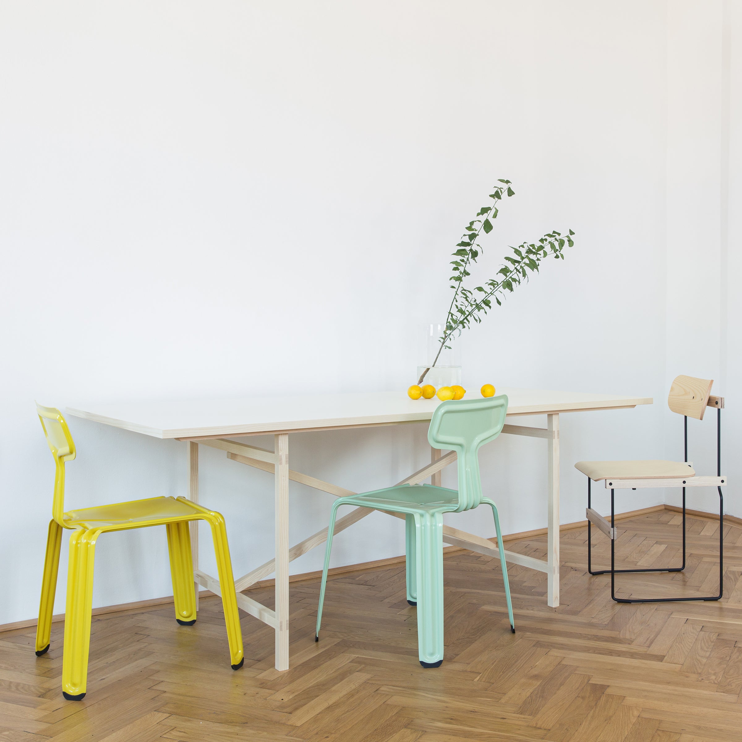The dining area showcases a Moormann Egon wooden table, accompanied by three mismatched chairs in yellow, mint green, and natural wood. A glass vase with green branches and lemons embellishes the table against a light wooden herringbone floor.