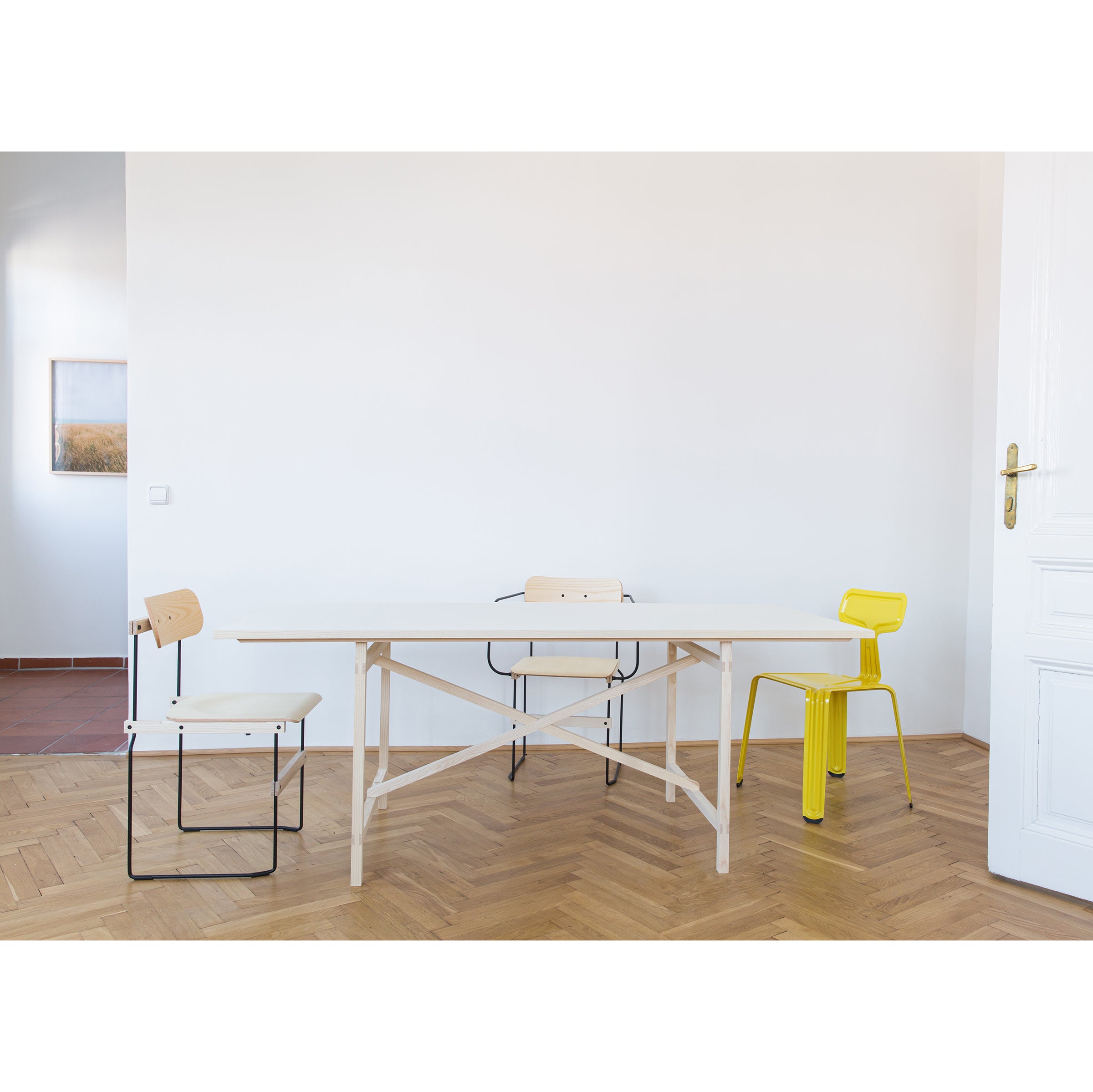 In a minimalist room with light wooden flooring and white walls, the Moormann Egon collection features a Franz Volhard-inspired, metal-free white table surrounded by three distinct chairs: black, natural wood, and bright yellow. A small framed picture hangs on the wall in the background.