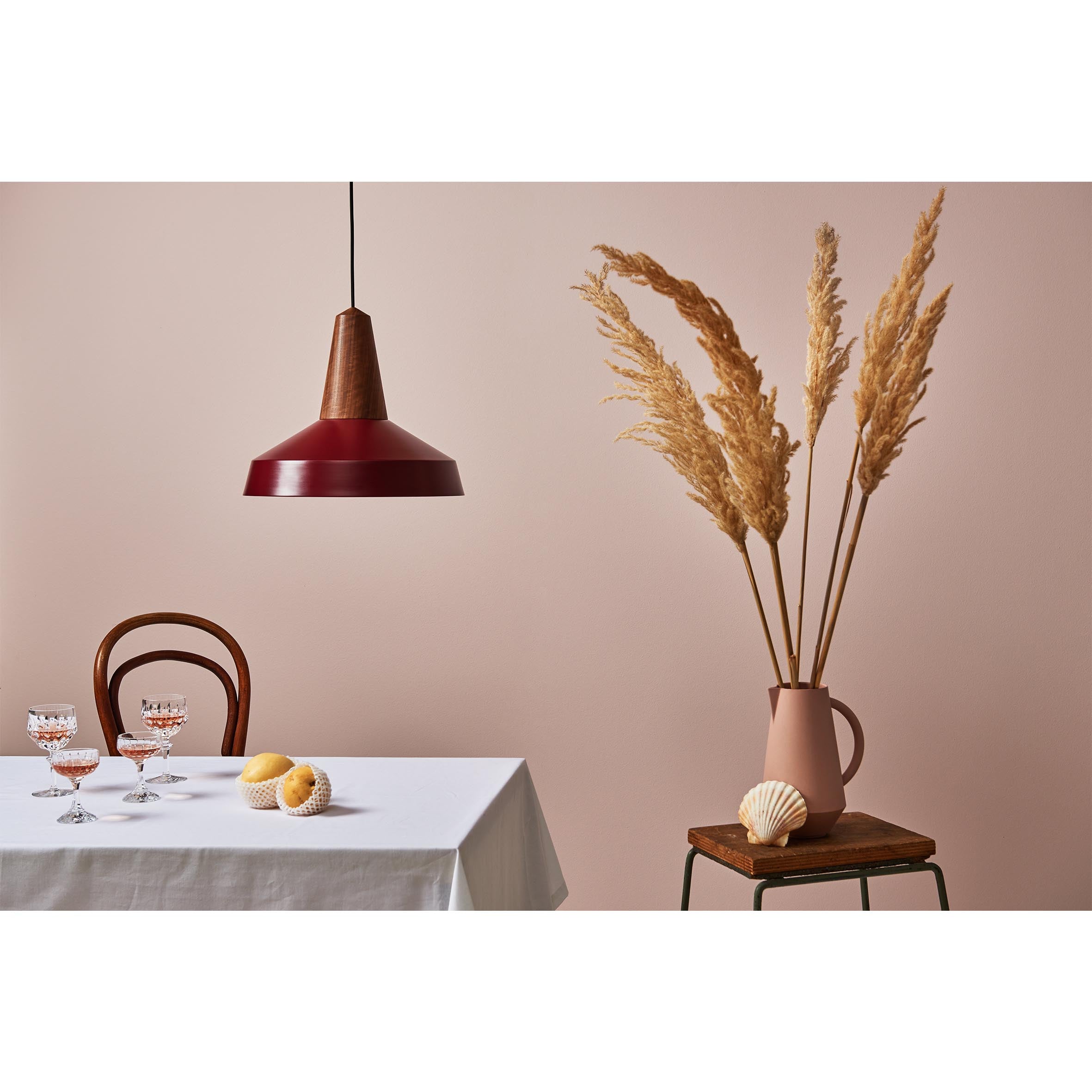 A minimalist dining setup features a white tablecloth, crystal glasses, and shells. Beneath the Schneid Eikon Circus Burgundy pendant light is a wooden chair. A stool holds a vase with pampas grass against a beige wall, crafting a contemporary expression.