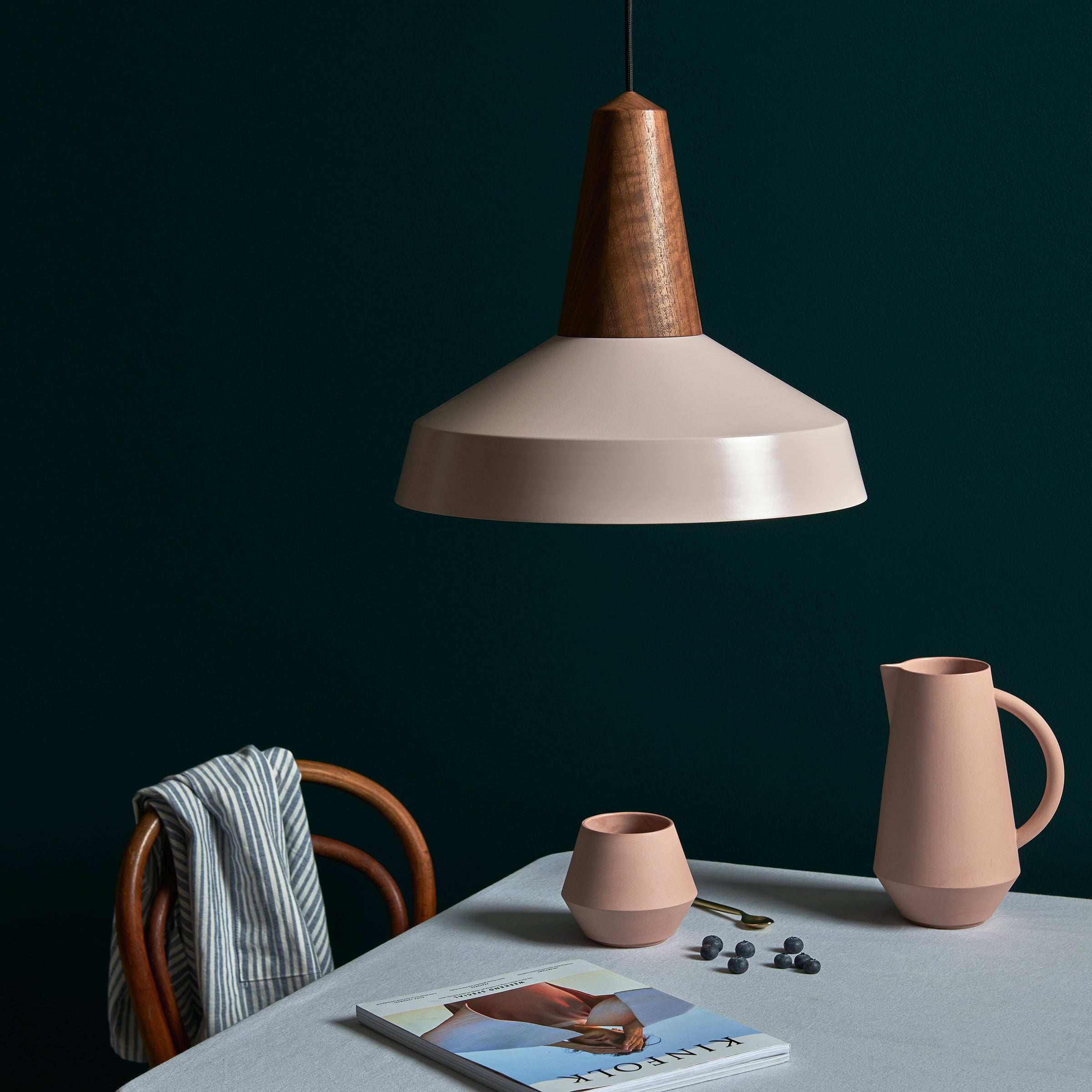 A minimalist scene shows a wooden chair, striped cloth, and a table with a pitcher, cup, scattered almonds, and a magazine. An Eikon Circus Pale Rose pendant lamp from Schneid glows overhead, set against a dark green backdrop with contemporary expression.