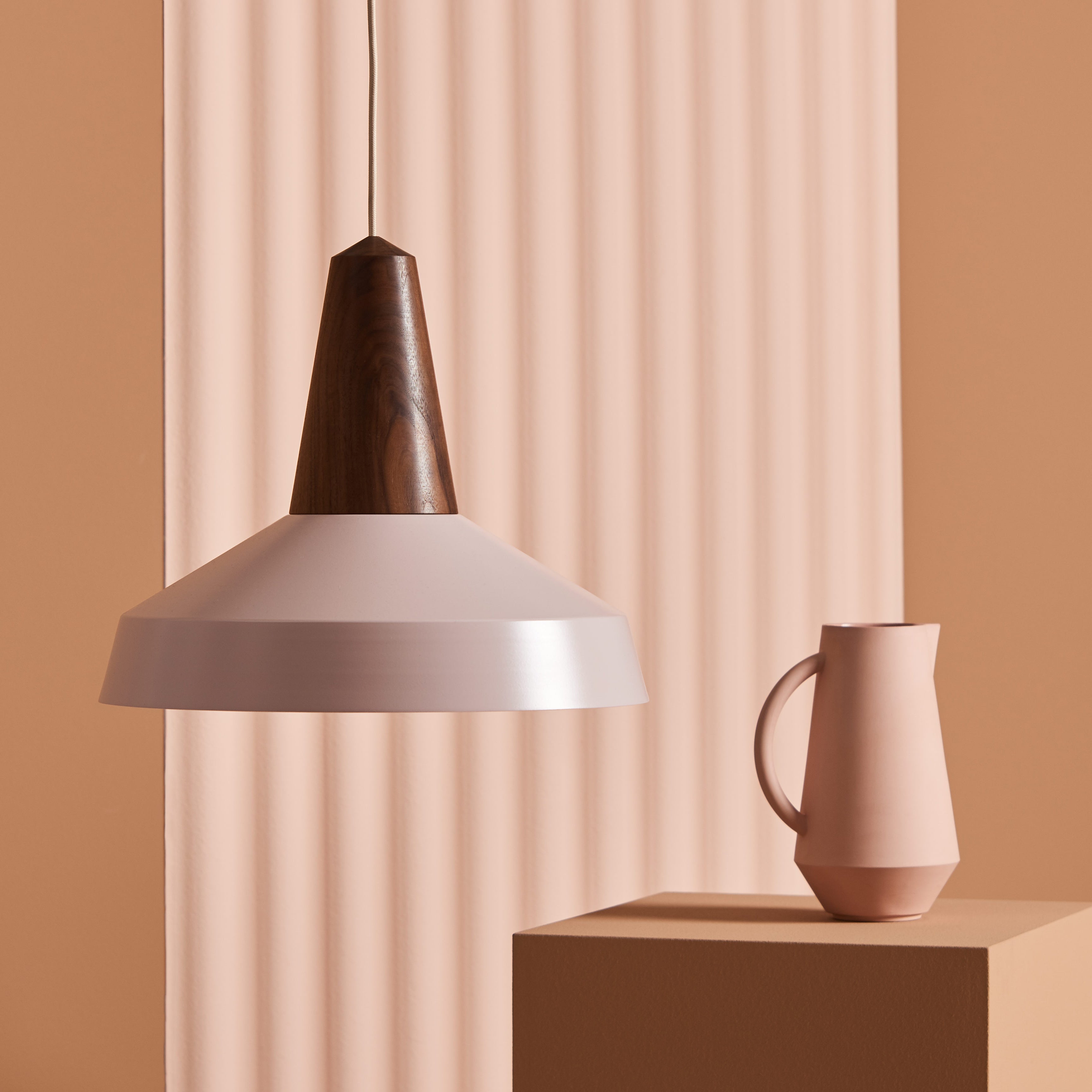 An Eikon Circus Pale Rose pendant lamp by Schneid, featuring a wooden top and white shade, hangs beside a matte pink ceramic pitcher on a beige pedestal. Vertical ridges in matching beige form the background, enhancing the contemporary vibe of the space.