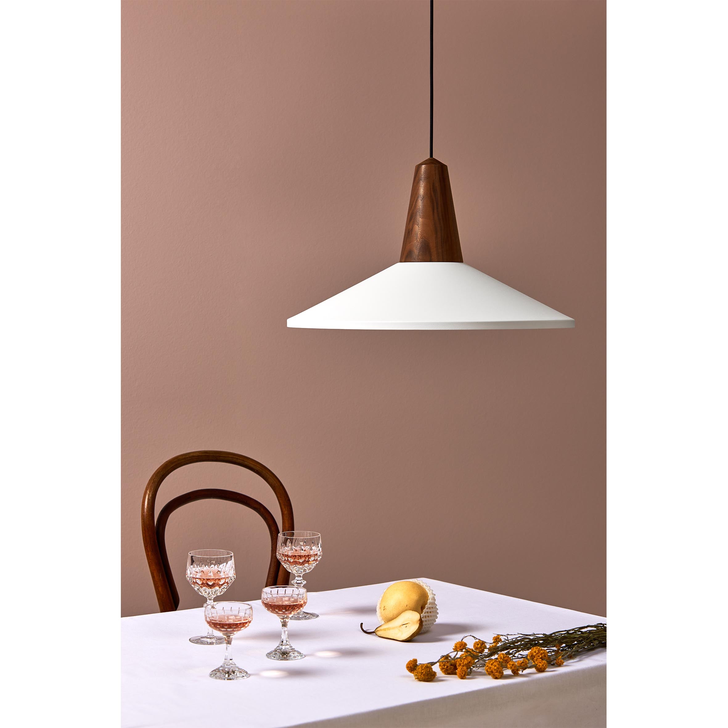 A stylish interior scene features a wooden chair, a white tablecloth with three glasses of pink drink, a yellow pear, and dried flowers. Above, Schneids Eikon Shell White pendant light in certified wood with a white shade and dark wood detail is suspended.