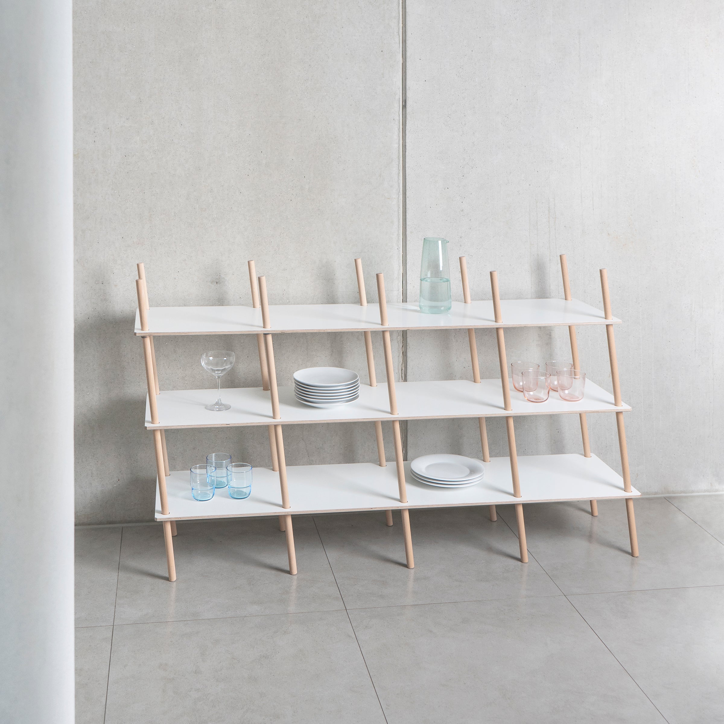 The Es Sideboard by Moormann is a minimalist shelving unit featuring white shelf boards with light birch plywood supports. Against a gray wall, it precisely holds plates, glasses, and a carafe while exuding a clean, modern aesthetic.