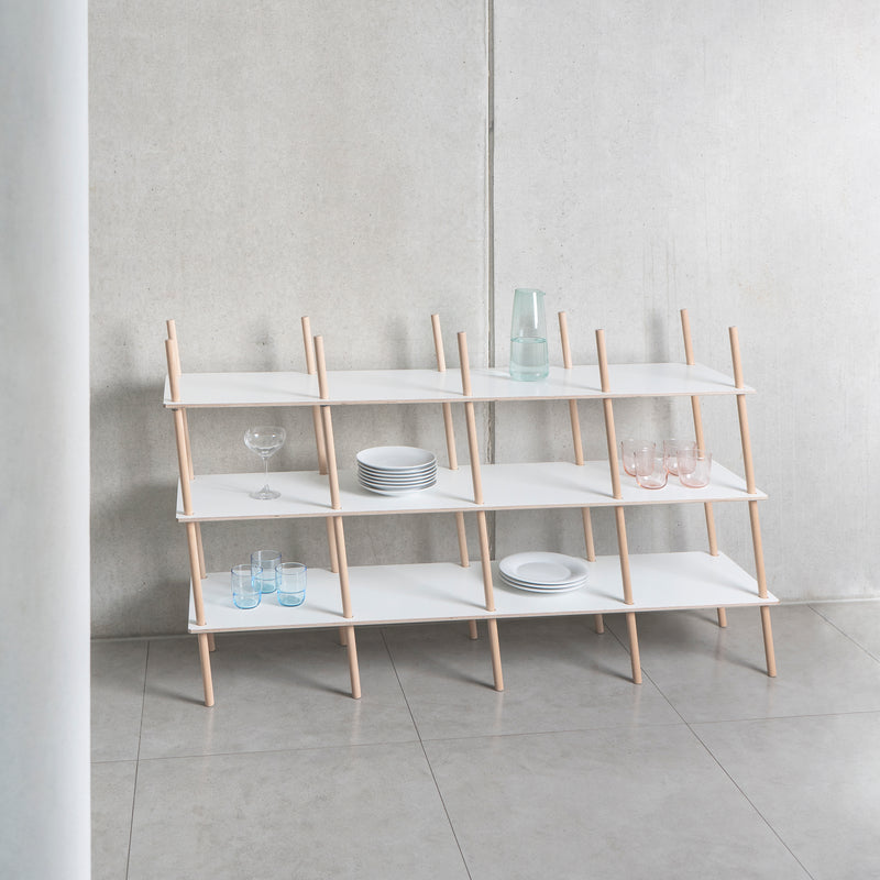 The Es Sideboard by Moormann is a minimalist shelving unit featuring white shelf boards with light birch plywood supports. Against a gray wall, it precisely holds plates, glasses, and a carafe while exuding a clean, modern aesthetic.