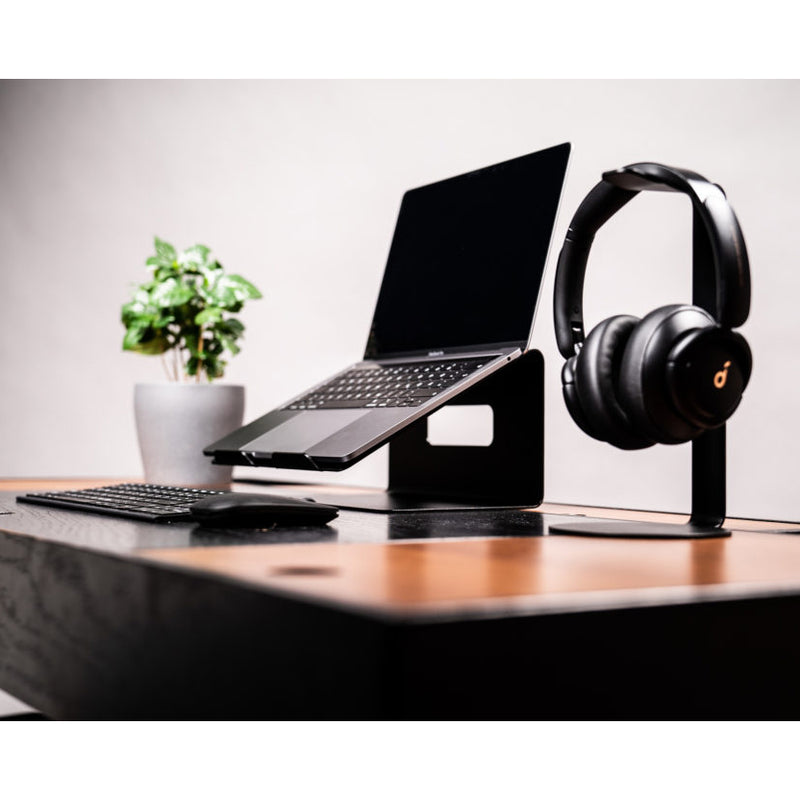 A sleek workspace includes an Echtstahl Laptop Stand holding a laptop, paired with a wireless keyboard and mouse. Headphones rest on their stand, while a small potted plant adds greenery. This setup sits on a polished wooden desk featuring a modern minimalist design and quality steel accents.