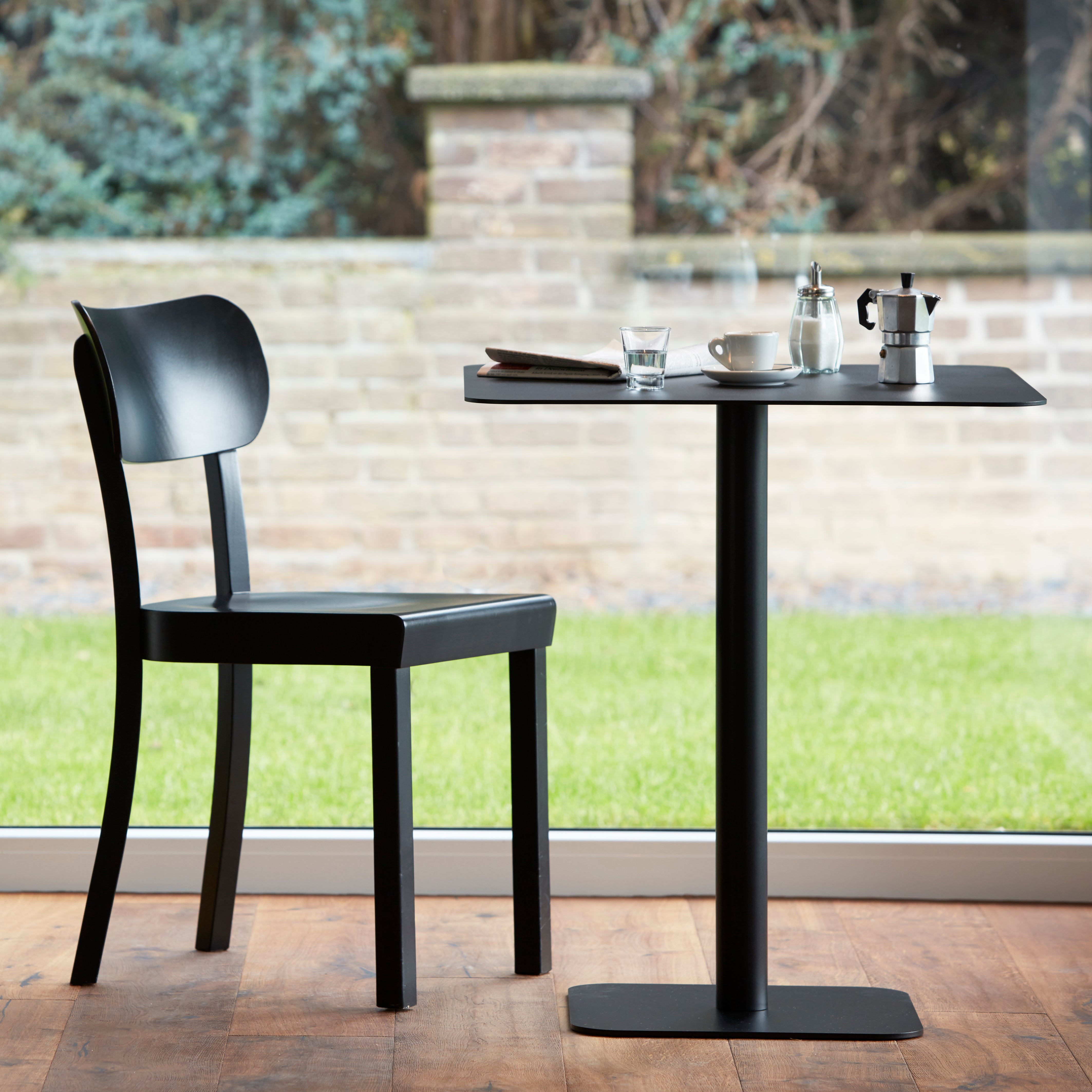 The chic dining area features the Bistro Table 123 from Echtstahl by Thomas Schnur, a square table made of powder-coated steel with a black chair. On it sit a cup, glass, small coffee pot, and saucer. Positioned near a large window, the setup overlooks a grassy lawn and stone wall.