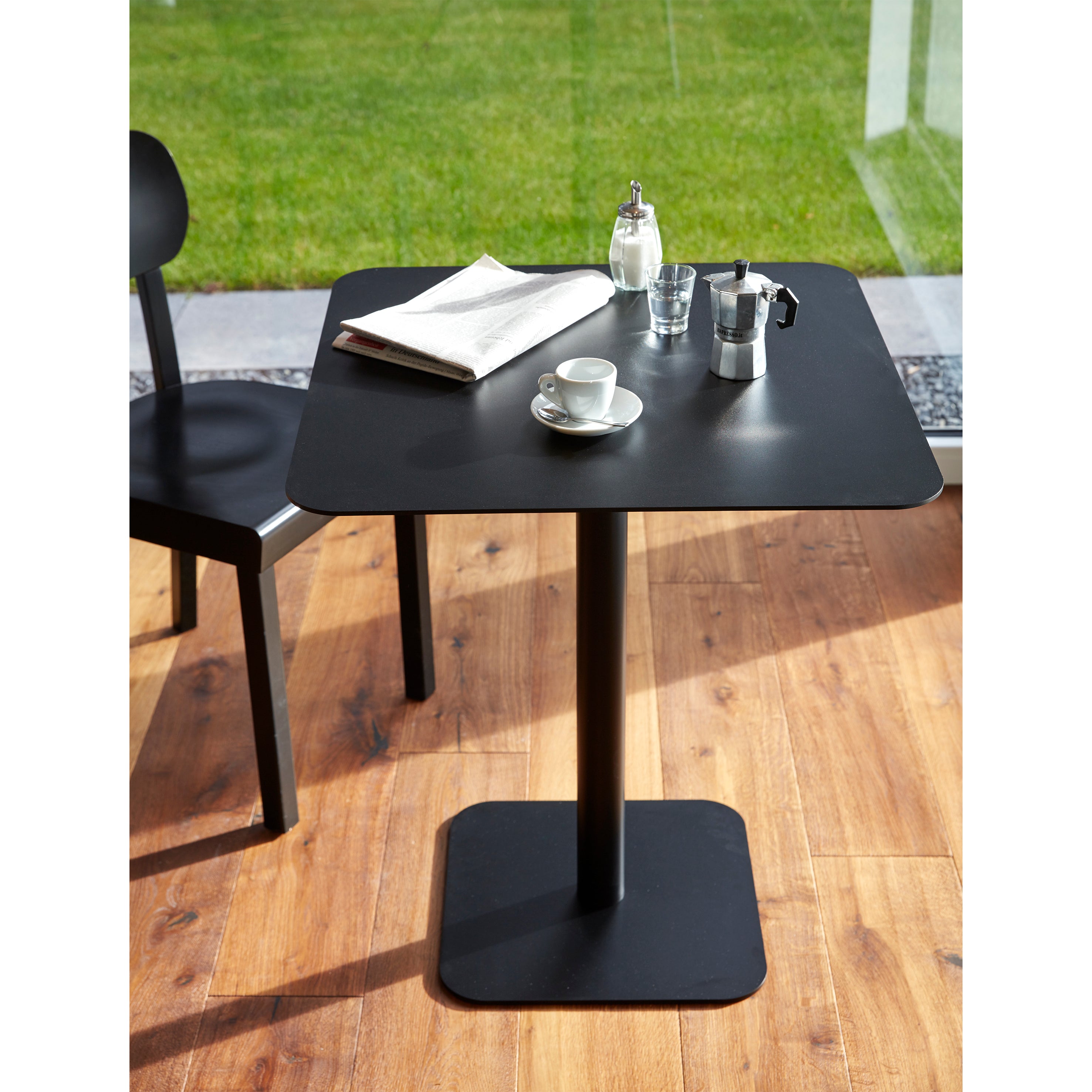 Placed on a wooden floor, the sleek black Bistro Table 123 from Echtstahl, designed by Thomas Schnur, supports a newspaper, coffee cup, small metal coffee maker, and sugar dispenser. A black chair is partially visible nearby while large windows offer a view of the green lawn outside.