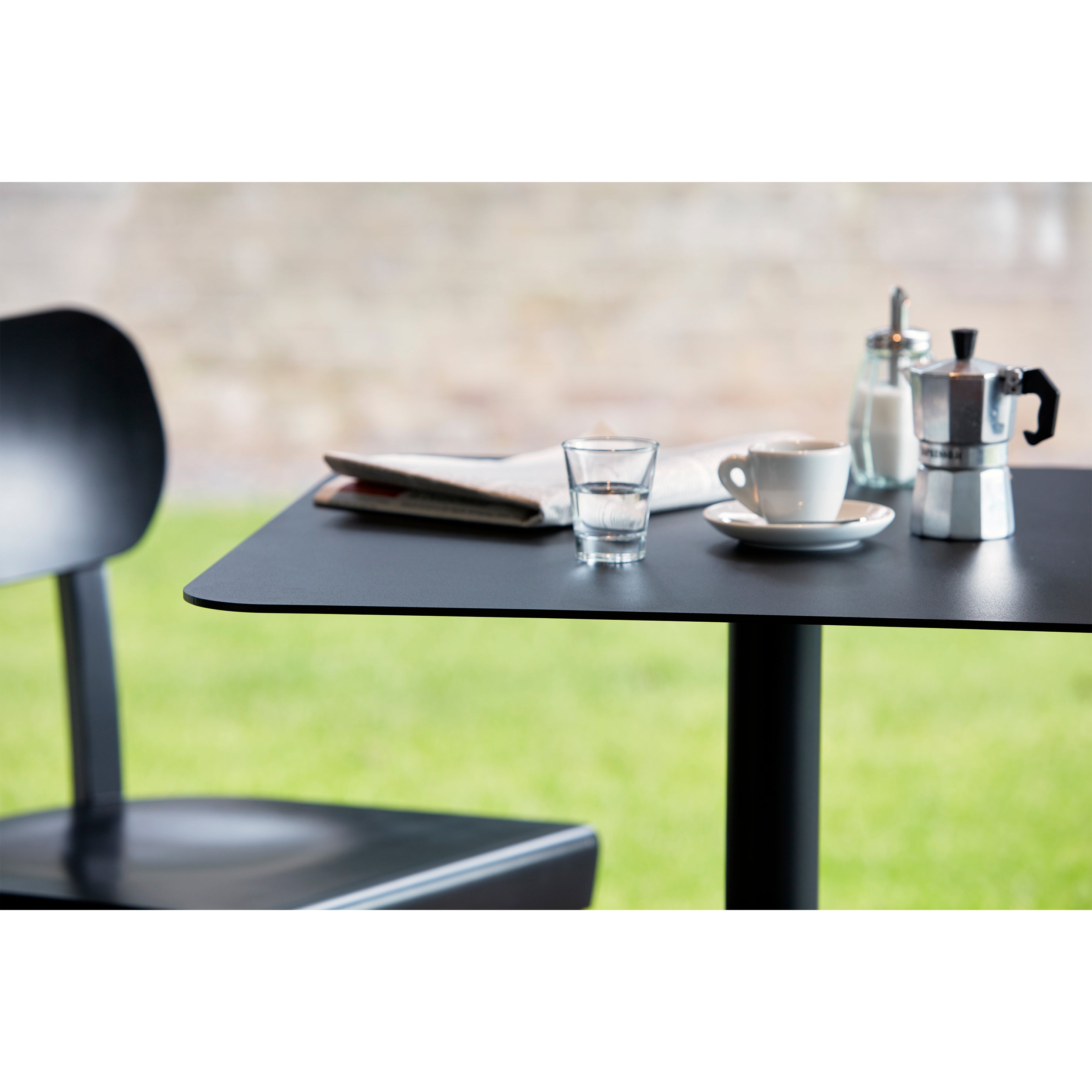 A sleek black Bistro Table 123 from Echtstahl serves a glass of water, a cup of coffee on a saucer, a moka pot, and a sugar dispenser. Nearby sits a folded newspaper. A powder-coated steel chair is partially visible against the blurred green lawn and brick wall backdrop.