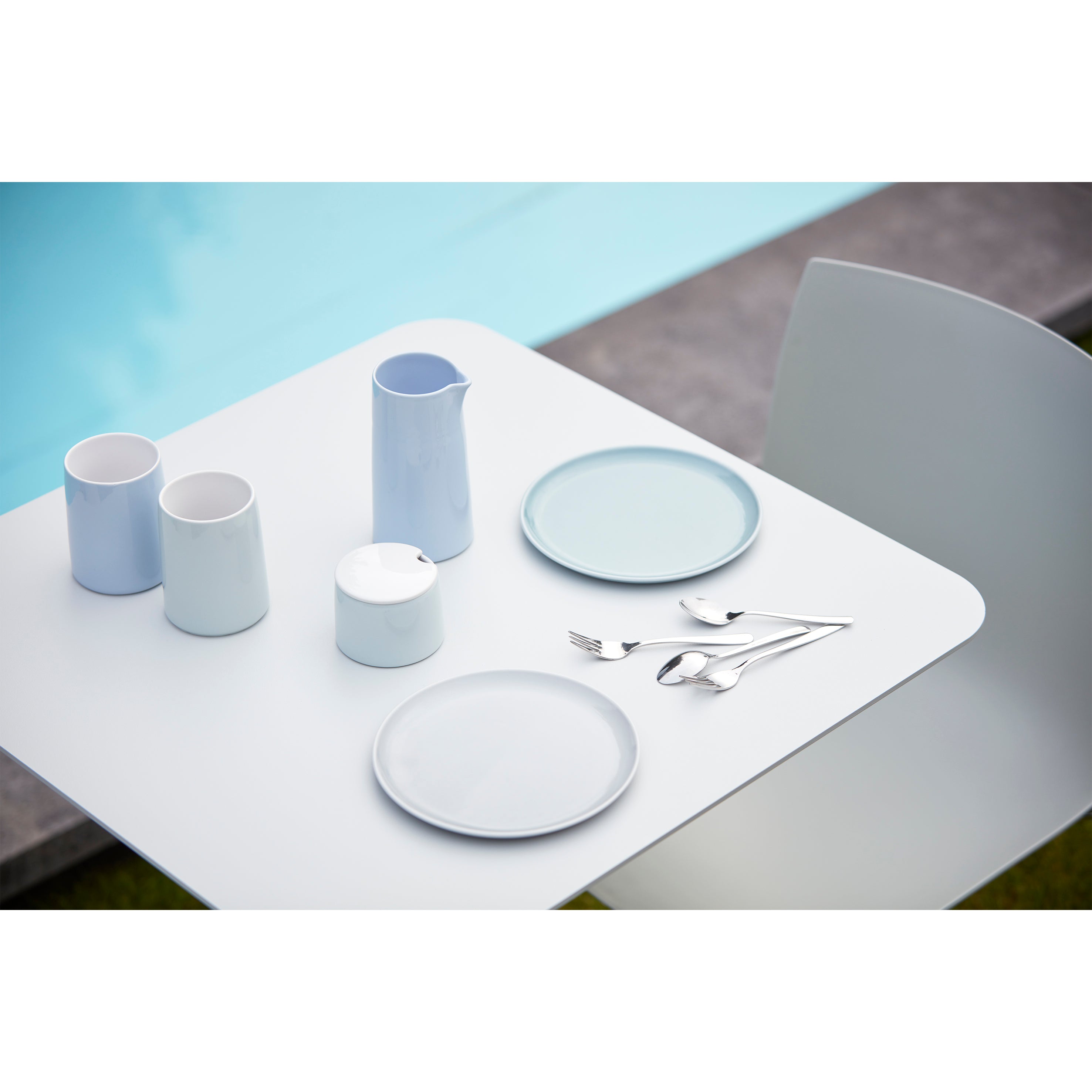A minimalist outdoor dining scene features the sleek Bistro Table 123 by Echtstahl in powder-coated steel. The white table by the pool displays two light blue plates, silverware, a pitcher, and mugs—all epitomizing clean modern style. A single white chair is partially visible.
