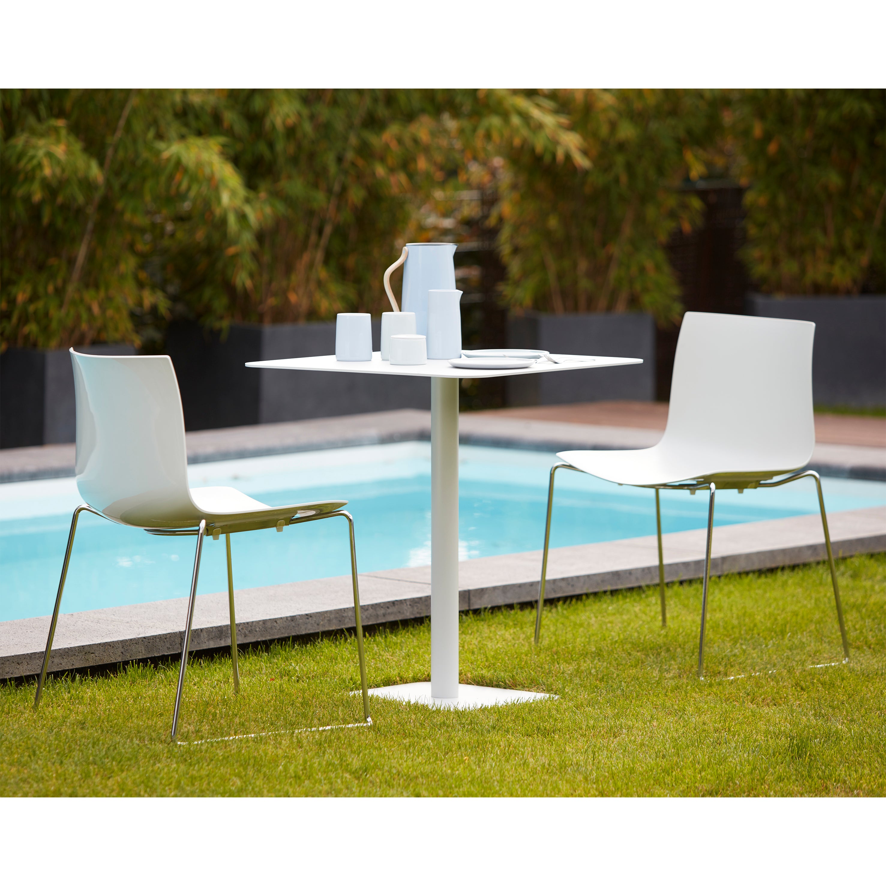 Two sleek white chairs and the Bistro Table 123 by Echtstahl, designed by Thomas Schnur from powder-coated steel, sit near a pool. The table holds a white teapot, cups, and a plate. Lush greenery in the background completes this serene outdoor setting.