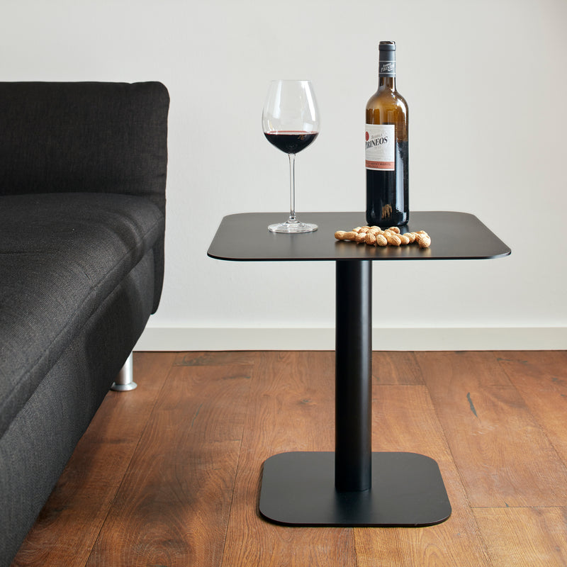 The Side Table 123 from Echtstahl, a small black table made of powder-coated steel, holds a bottle of red wine, a wine glass, and nuts next to a black sofa on wooden flooring.