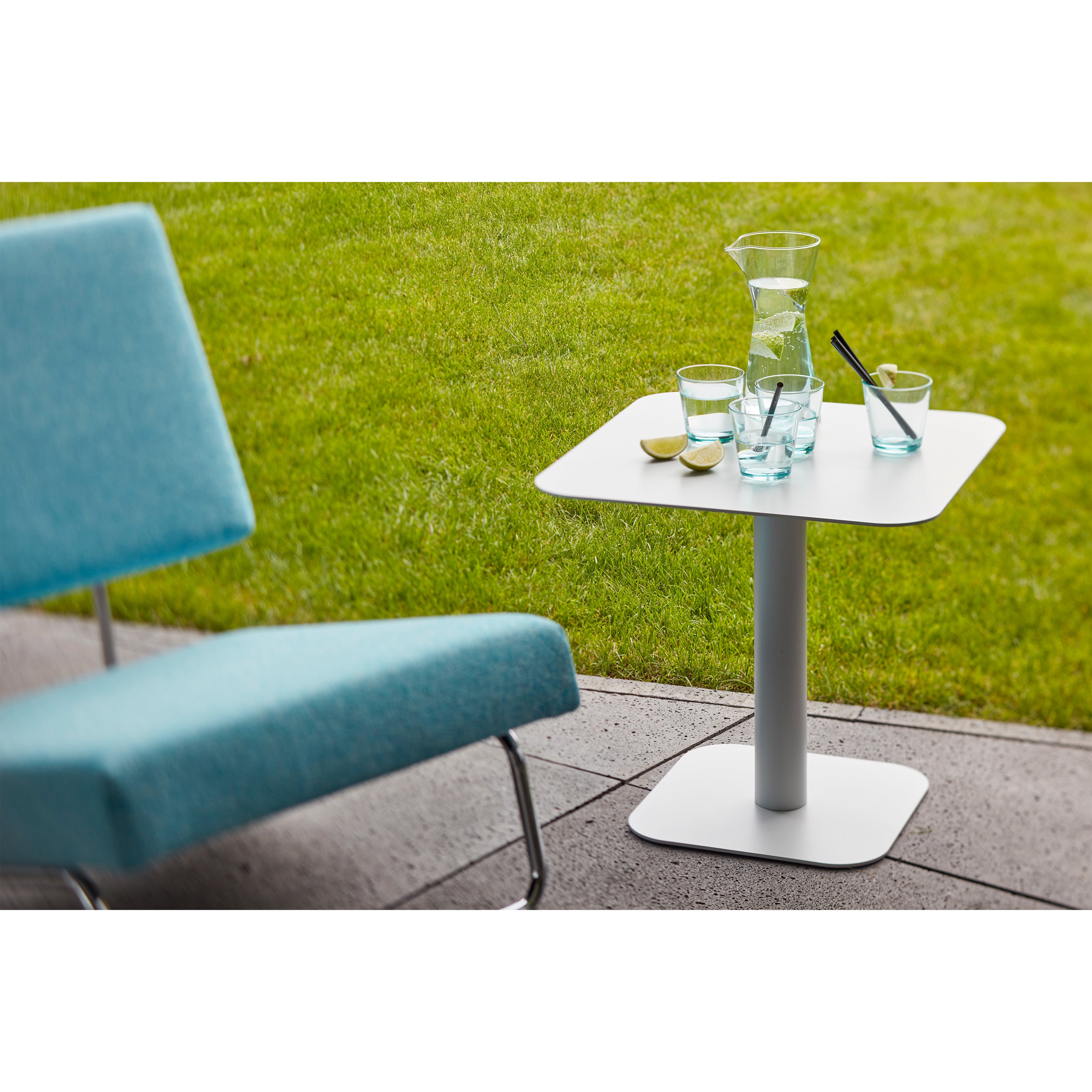 The Side Table 123 from Echtstahl, a small white square piece made of powder-coated steel, adorns the patio. It holds a glass carafe with two glasses of water, a straw, and lemon slices. A light blue chair nearby complements the vibrant green grass in the background.