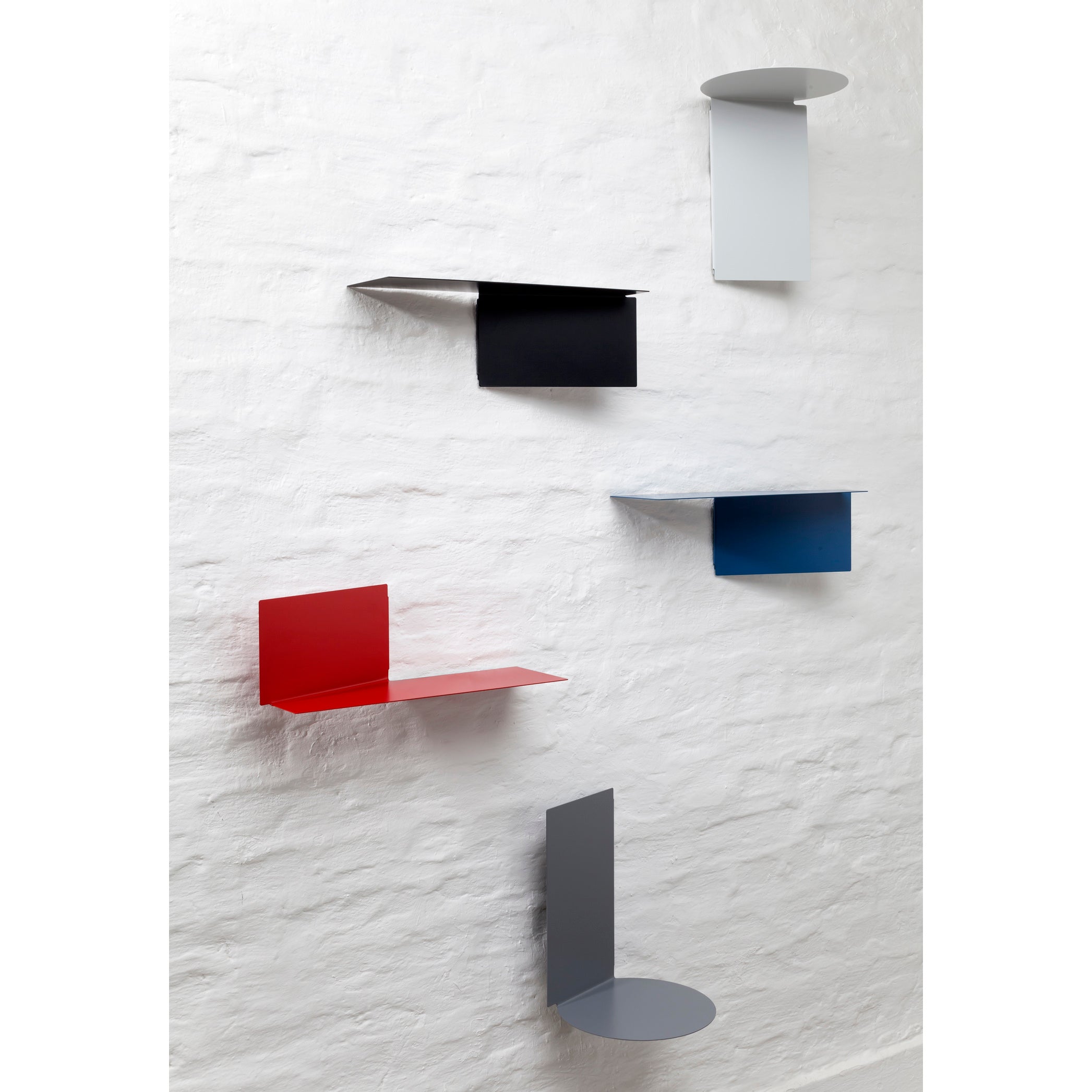 A set of Plateau rectangular shelves from Echtstahl is mounted on a white textured wall. Made of powder-coated steel, these colorful minimalist shelves come in red, black, gray, and blue, featuring thin panels with unique geometric designs for a chic touch.