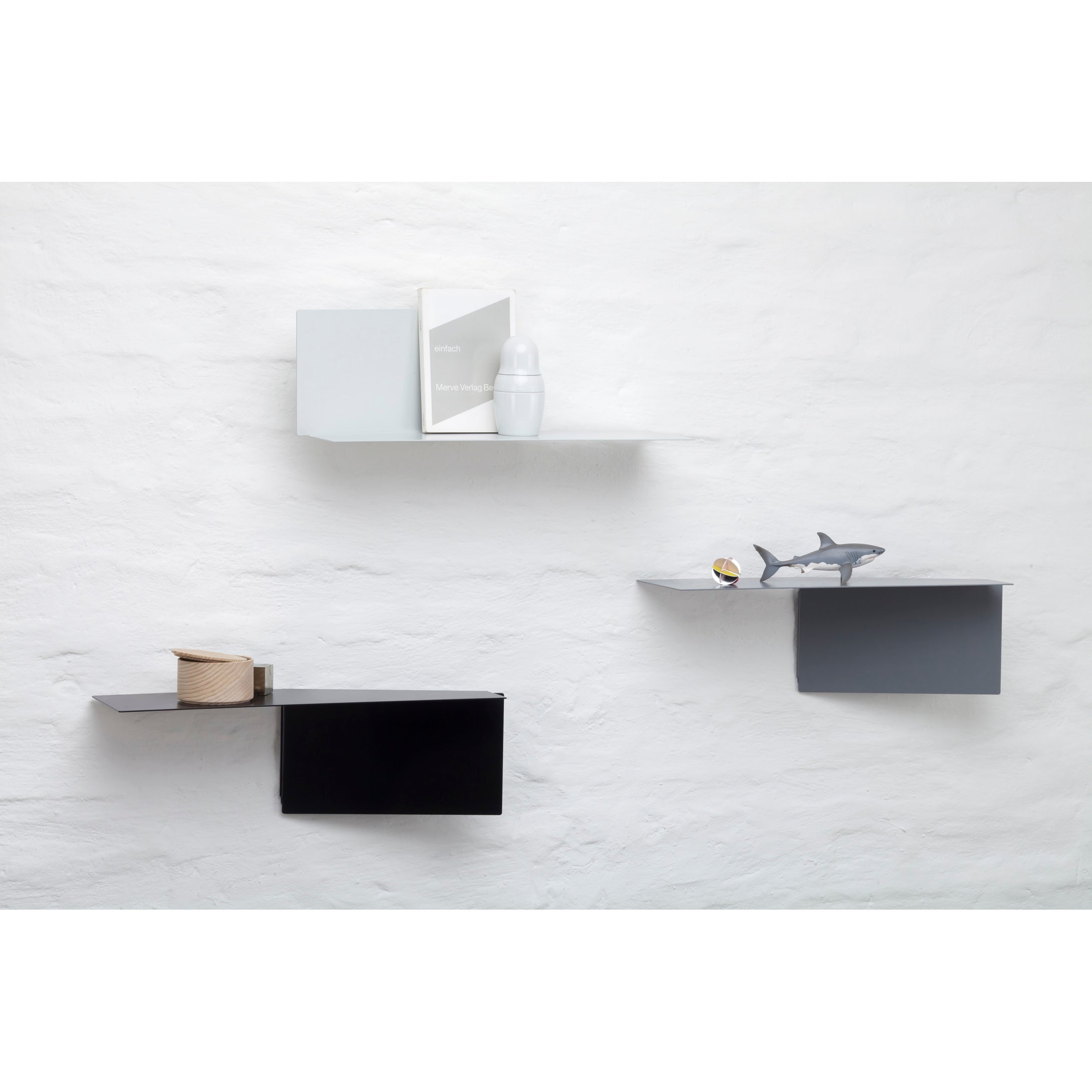 A minimalist white wall showcases sophisticated shelving: Echtstahls powder-coated steel Plateau rectangular shelf holds a book and container, a gray version holds a shark figurine and sphere, while the black shelf supports a wooden roll.