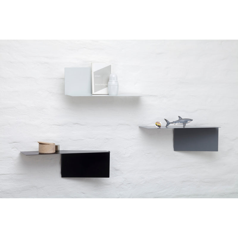 A minimalist white wall showcases sophisticated shelving: Echtstahls powder-coated steel Plateau rectangular shelf holds a book and container, a gray version holds a shark figurine and sphere, while the black shelf supports a wooden roll.