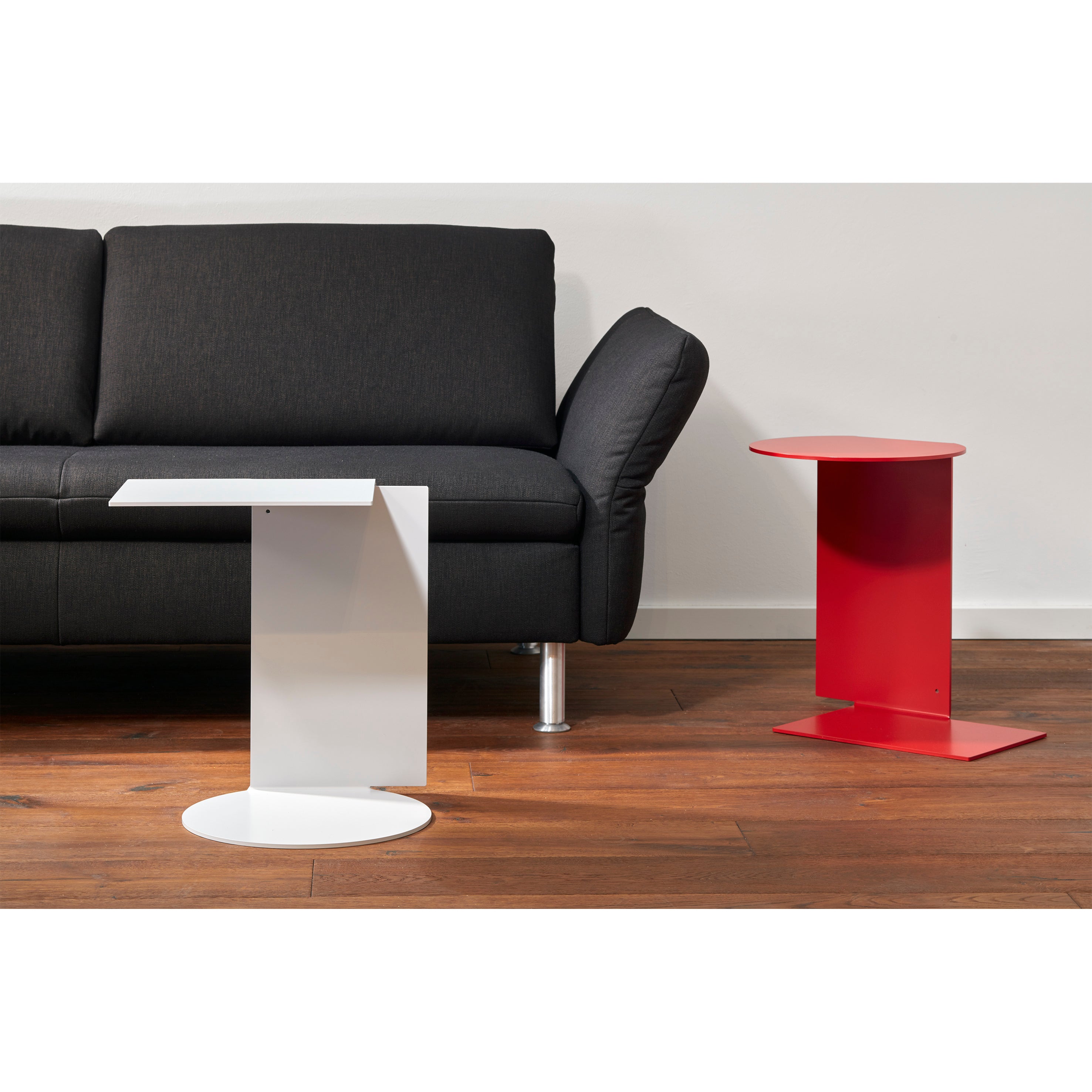 A modern living room features a black sofa on wooden flooring. Beside it is a unique white side table with a geometric design and the Echtstahl red Plateau side table, made of powder-coated steel. The plain white wall enhances this versatile setup.
