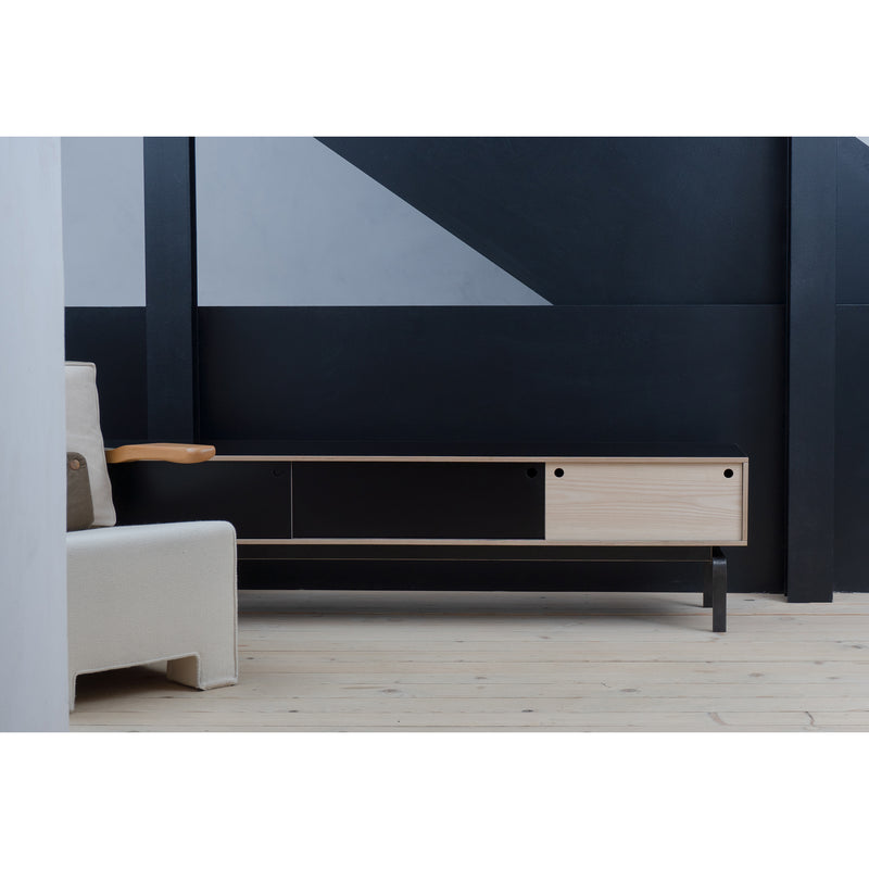 A minimalist living room showcases the Egal Lowboard by Moormann, with light wood and black elements set against a geometric black and gray wall. A beige sofa with wooden armrests is partially visible on the left, while light wooden flooring enhances the modern aesthetic.