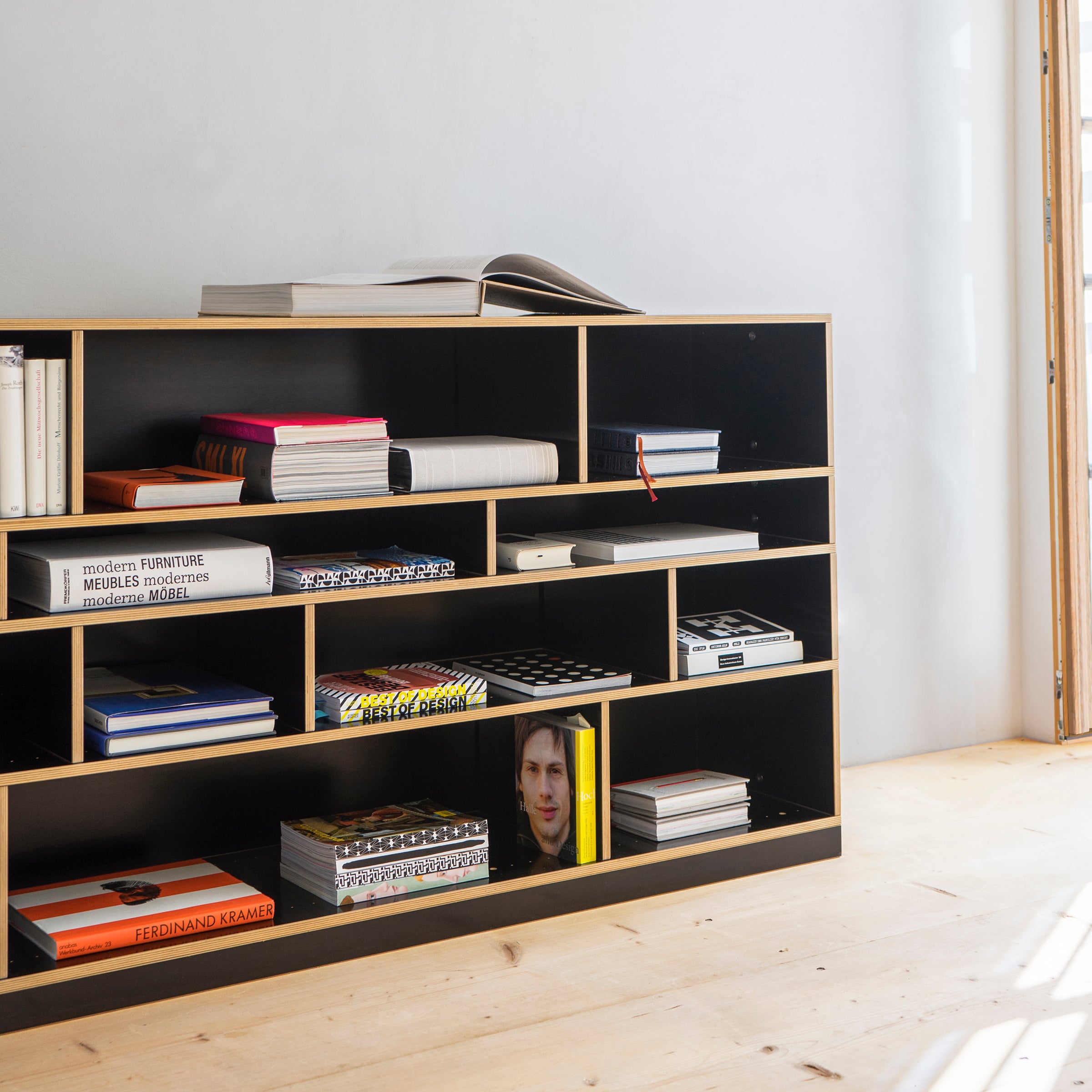 The Moormann Egal shelf system, in black, features open, customizable compartments filled with books and magazines. A bright yellow book with a face stands out. Light wood flooring and soft lighting create a cozy atmosphere, while an open book on the top shelf invites exploration.