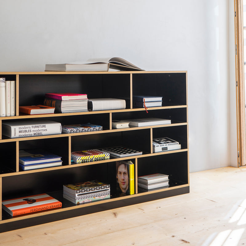 The Moormann Egal shelf system, in black, features open, customizable compartments filled with books and magazines. A bright yellow book with a face stands out. Light wood flooring and soft lighting create a cozy atmosphere, while an open book on the top shelf invites exploration.
