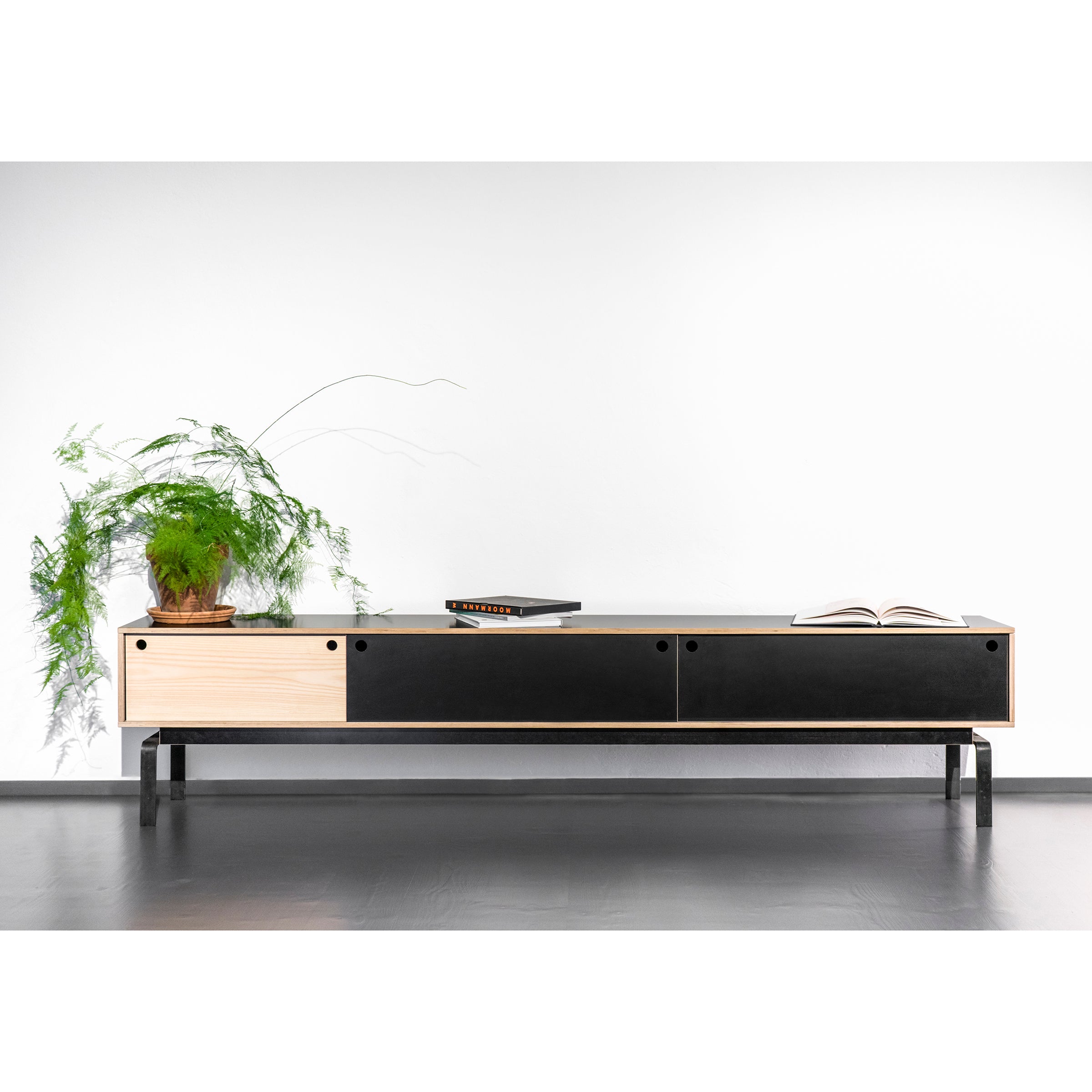 The Moormann Egal Lowboard, a modern black and wood sideboard on black legs, elegantly stands against a white wall. It showcases two sections with sleek doors and is adorned with a potted fern, a closed laptop, and an open book.
