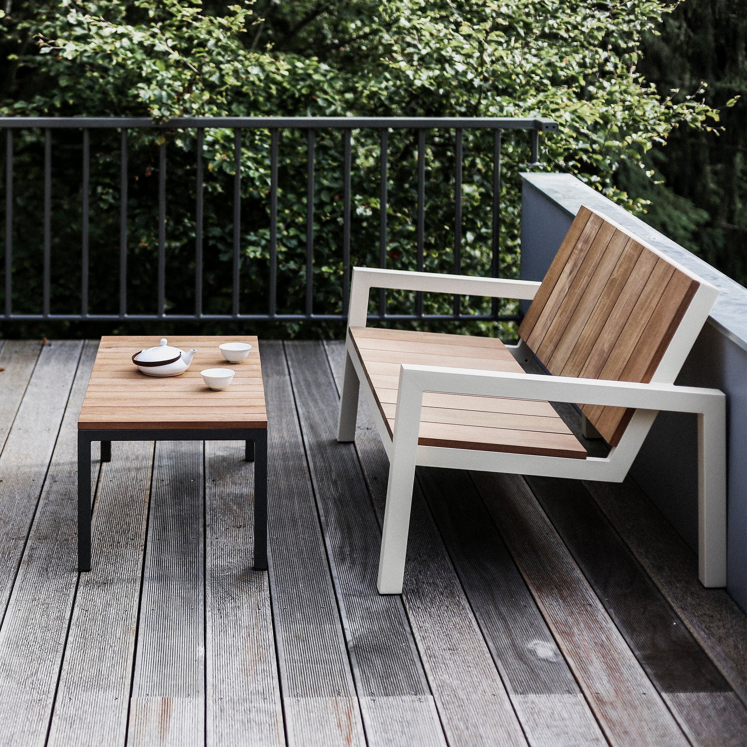 The Egoe Laurede series wooden patio set adorns the deck with a two-seater bench on a zinc-coated steel frame and a matching coffee table. A teapot and cups sit on the Laurede Low Table, amidst lush greenery, creating a peaceful atmosphere.