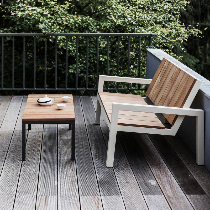 A modern outdoor patio features an Egoe Laurede Low Table and wooden bench set on a gray deck, with a tea set adorning the table. Beyond the zinc-coated steel black railing lies lush green foliage.