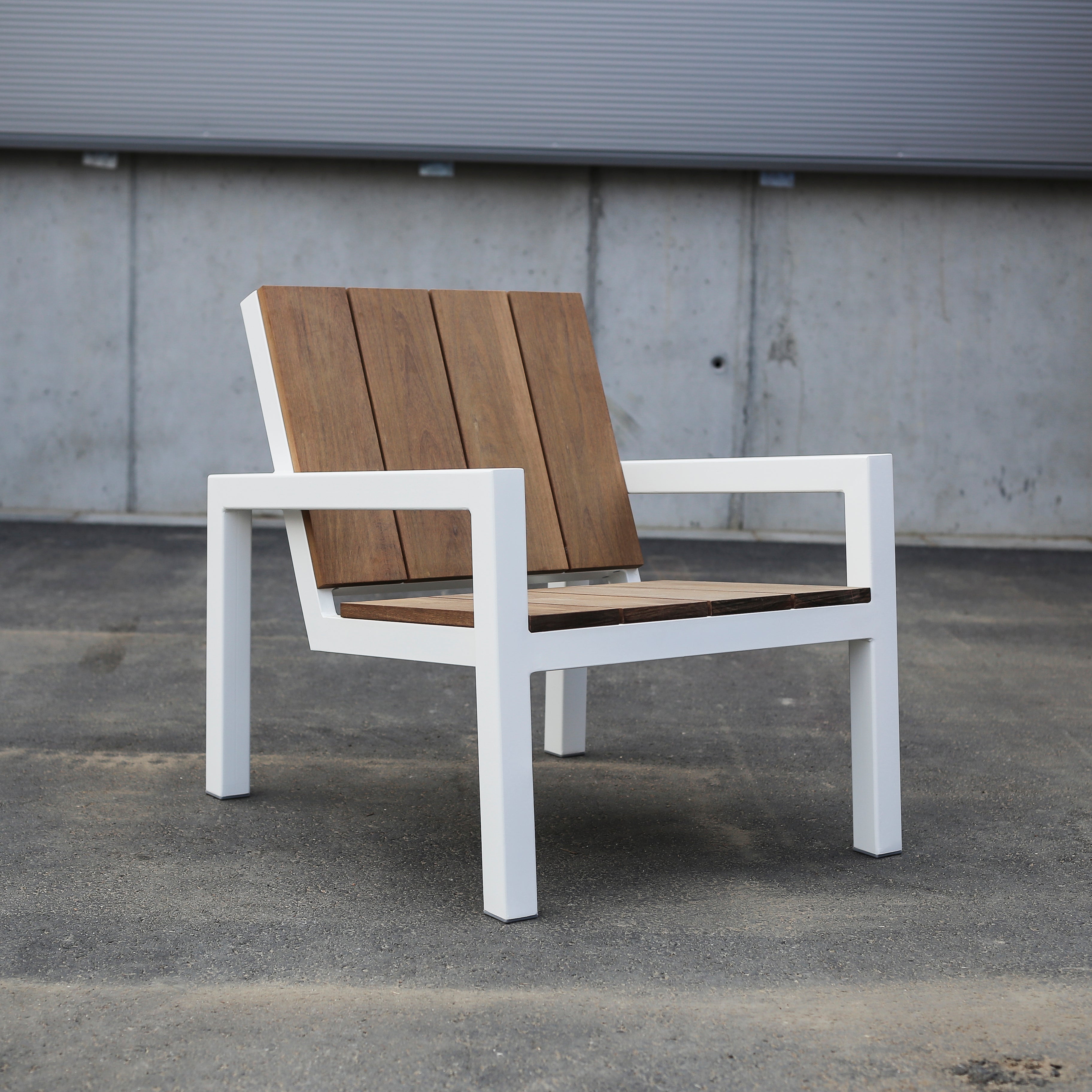 The Laurede Lounge Chair by Egoe showcases a sleek white metal frame with tropical wood slats for seat and backrest, elegantly set on concrete in front of a corrugated metal wall.