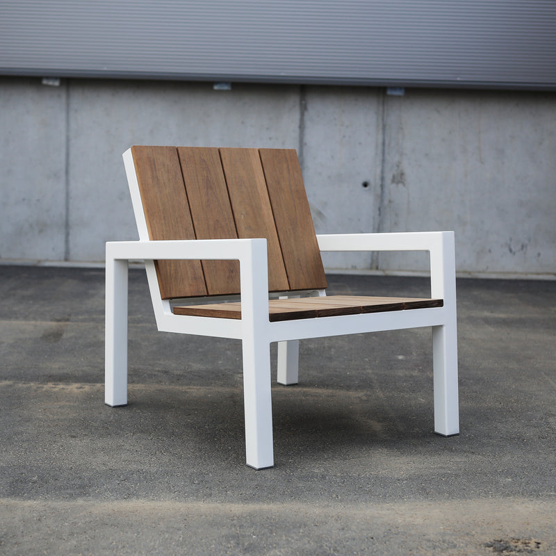 The Laurede Lounge Chair by Egoe showcases a sleek white metal frame with tropical wood slats for seat and backrest, elegantly set on concrete in front of a corrugated metal wall.