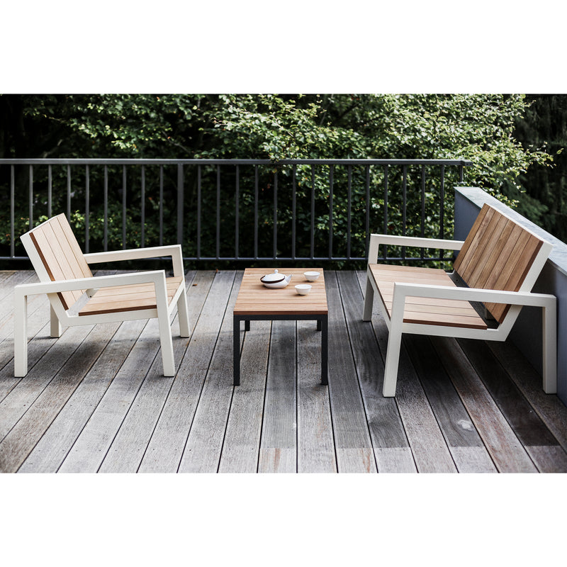 Two wooden chairs with white zinc-coated steel frames from the Laurede series by Egoe sit on a wooden deck. A Laurede Low Table crafted from tropical wood with a black metal frame stands between them, while lush green trees provide a picturesque backdrop.