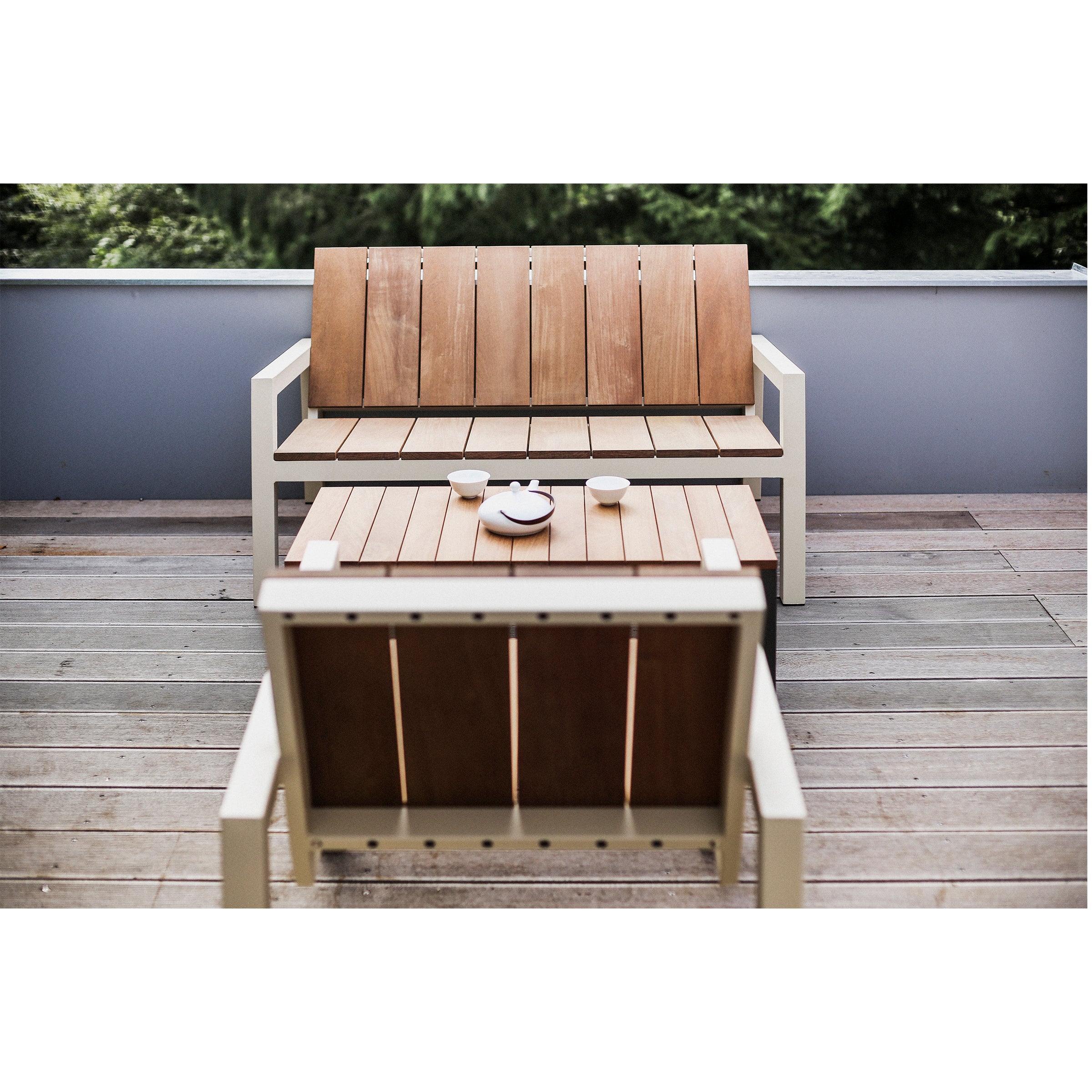 The Egoe Laurede wooden outdoor set beautifies the patio with a chair and bench around the Laurede Low Table, crafted from tropical wood, supporting a teapot and two cups. Green foliage thrives over a low wall in the background.