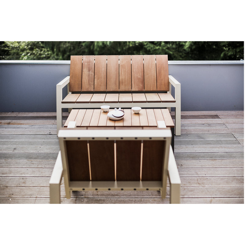 The Egoe Laurede wooden outdoor set beautifies the patio with a chair and bench around the Laurede Low Table, crafted from tropical wood, supporting a teapot and two cups. Green foliage thrives over a low wall in the background.