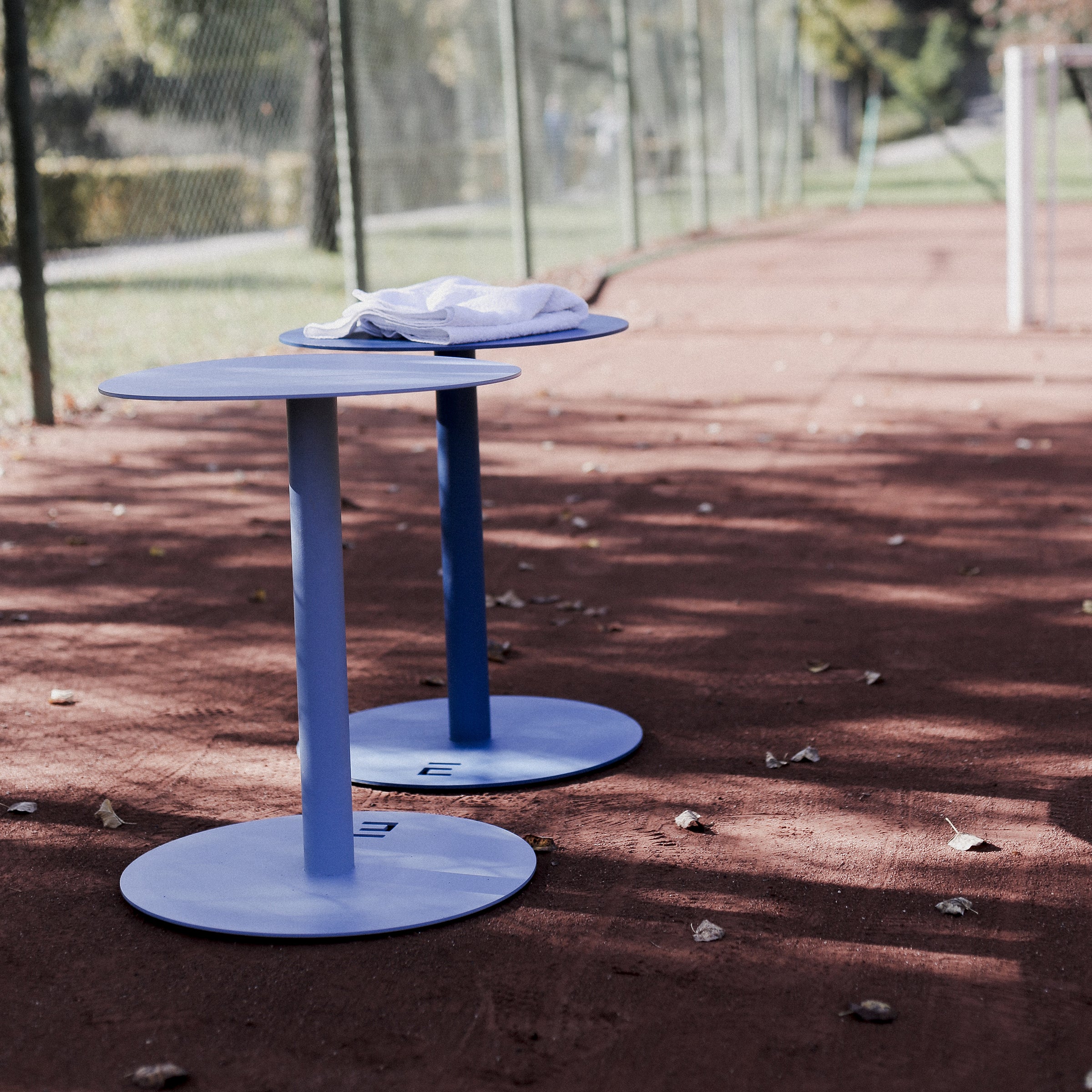 Two Egoe Spulka minimalist blue circular tables, featuring a durable all-metal body, sit on a red outdoor tennis court. Each is marked with a letter, and one has a white towel draped over it. Fencing and trees frame the sunny backdrop.